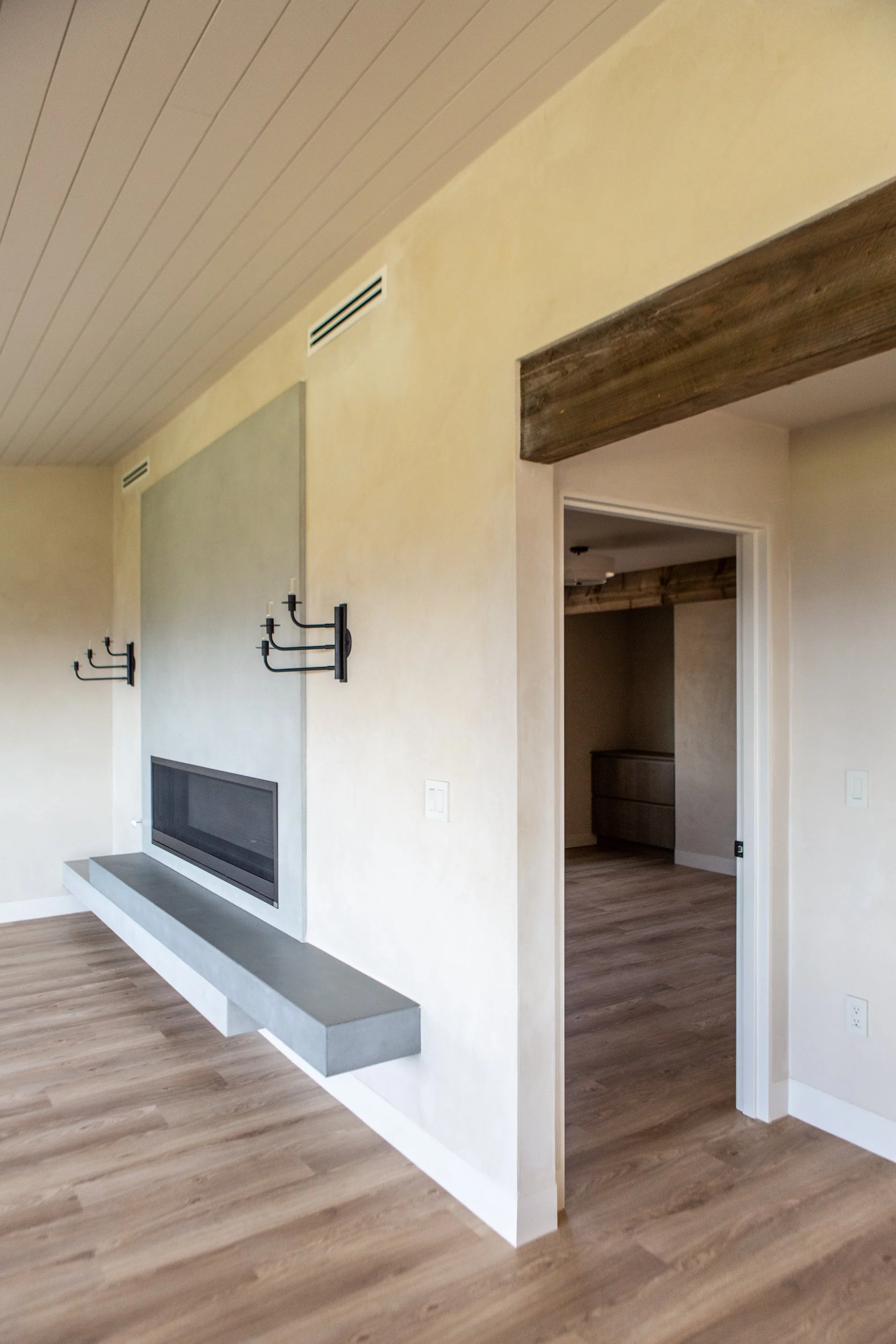 A modern living room with beige walls, wood flooring, a white ceiling with wood paneling, a fireplace with a gray concrete mantel, and black hooks on the wall. There is a doorway leading to another room with similar wood flooring and a wooden cabinet