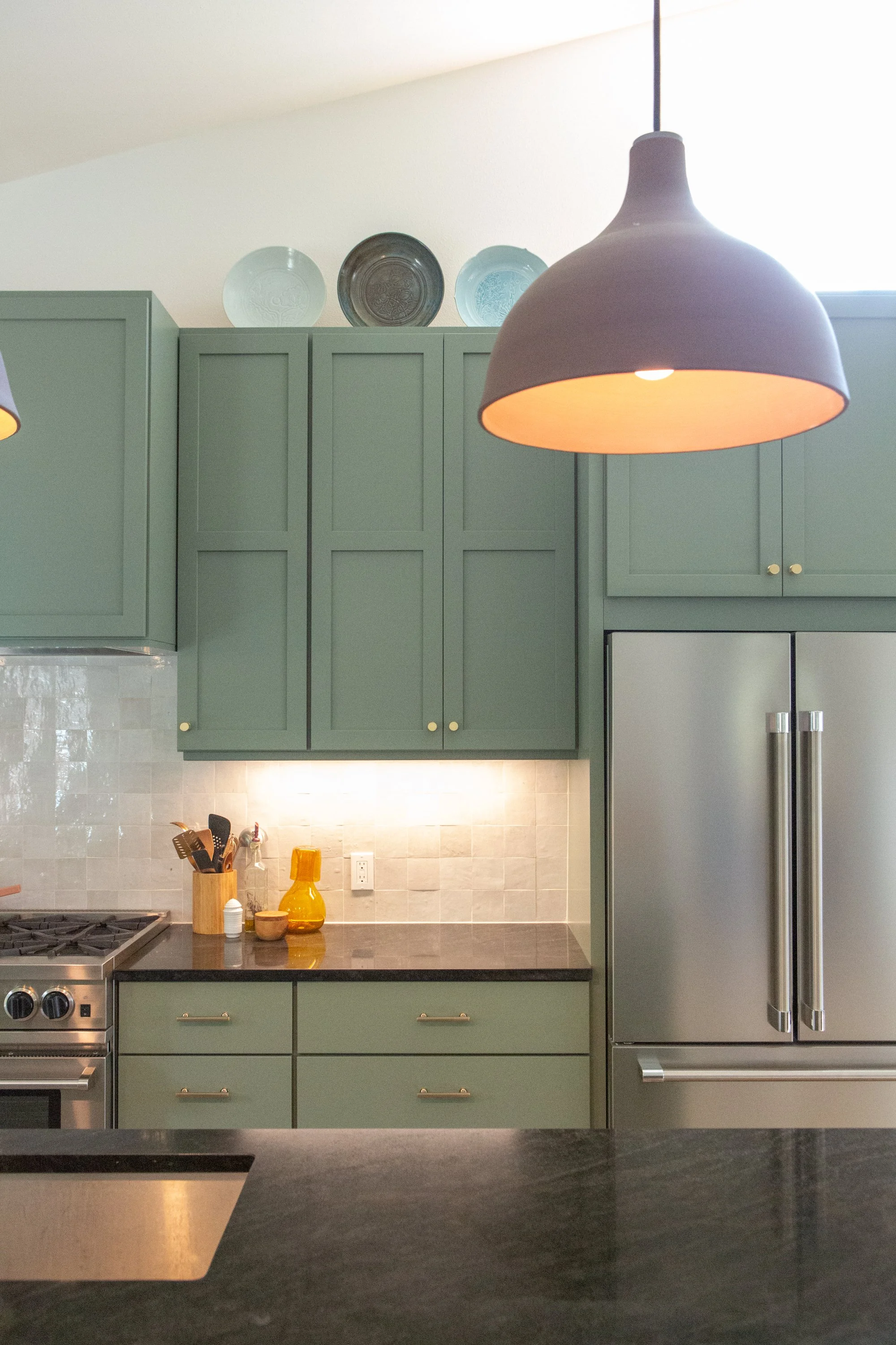 A modern kitchen with green cabinets, a black countertop, and stainless-steel appliances including a double-door refrigerator. Two black pendant lights hang from the ceiling, illuminating the space. Decorative plates are displayed on top of the cabin