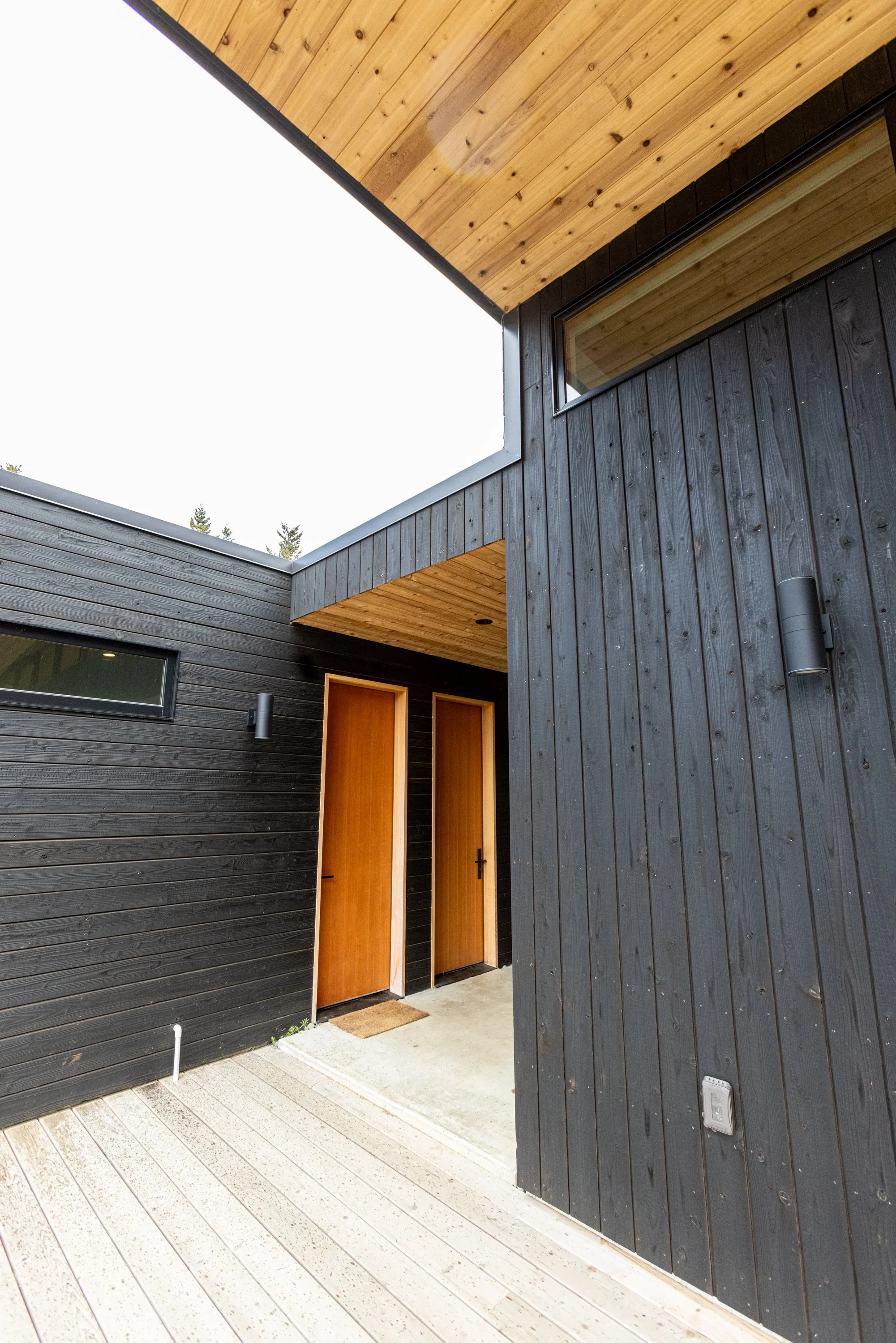 Exterior view of a modern house with black wooden siding, a large window, and a wooden front door, with a wooden ceiling above the entrance area and a light-colored deck in the foreground.