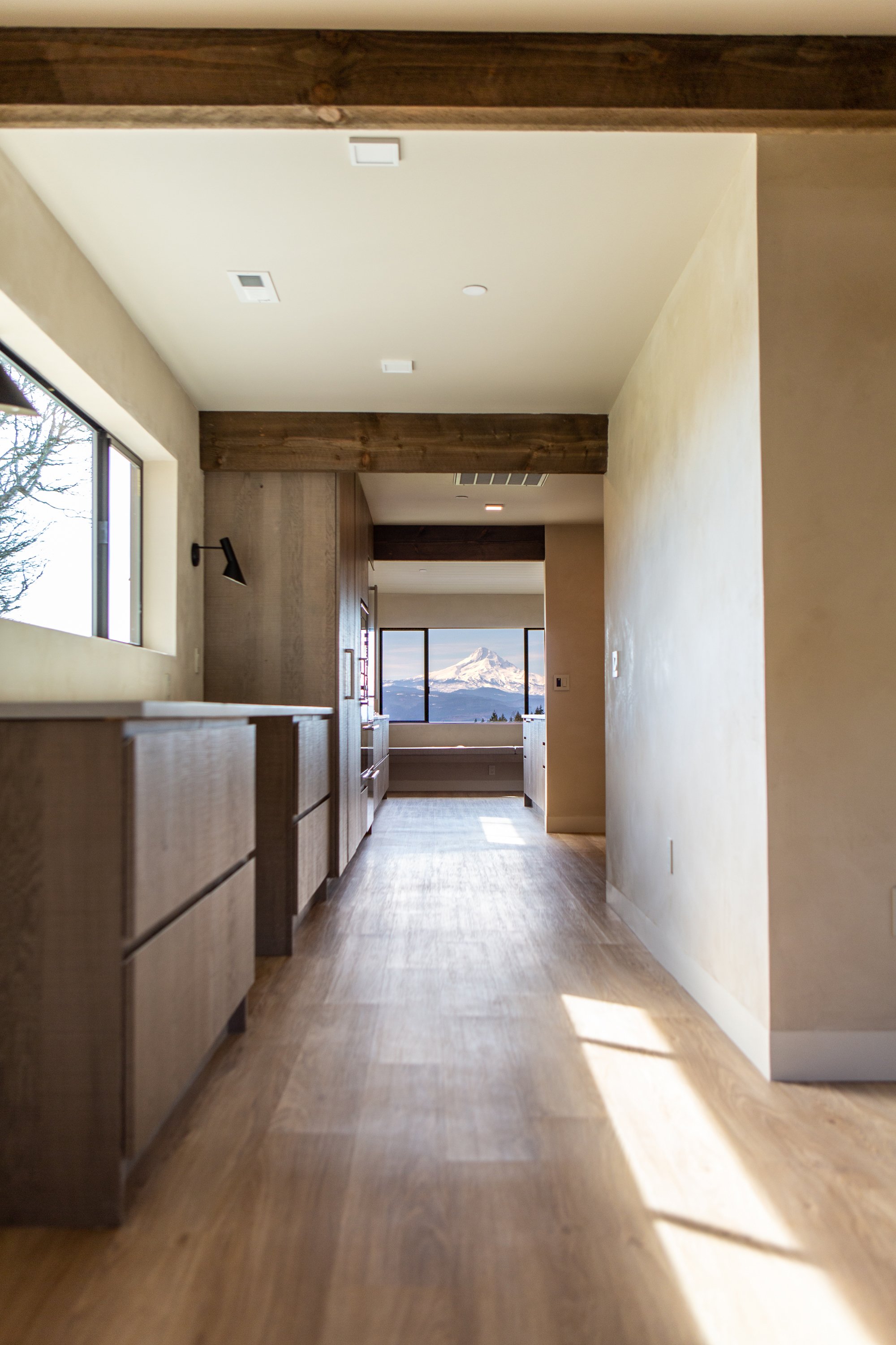 View through an interior corridor with wooden flooring, walls, and ceiling beams, leading to a window showing a snow-capped mountain landscape in the distance.