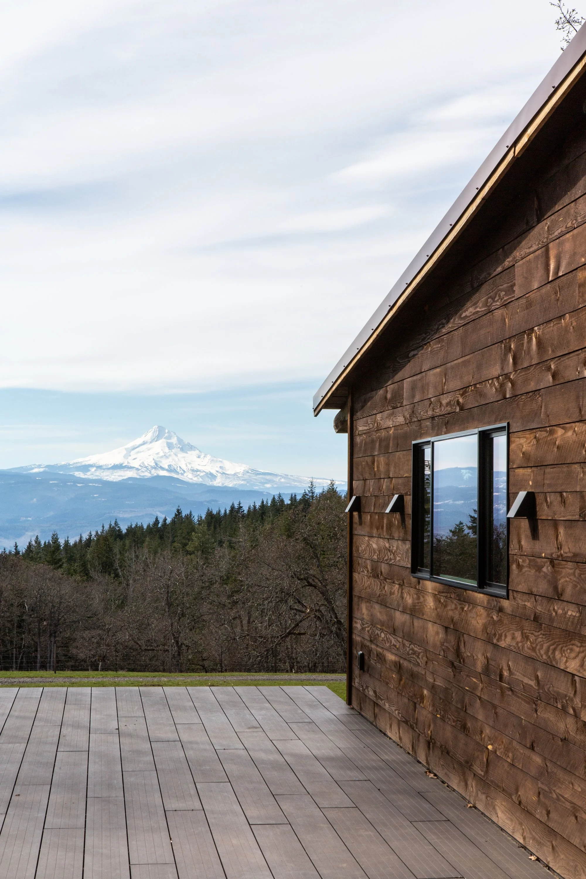 Partial view of a house with wood siding and a large window, overlooking a landscape with a forest, mountains, and a snow-capped peak in the background under a cloudy sky.