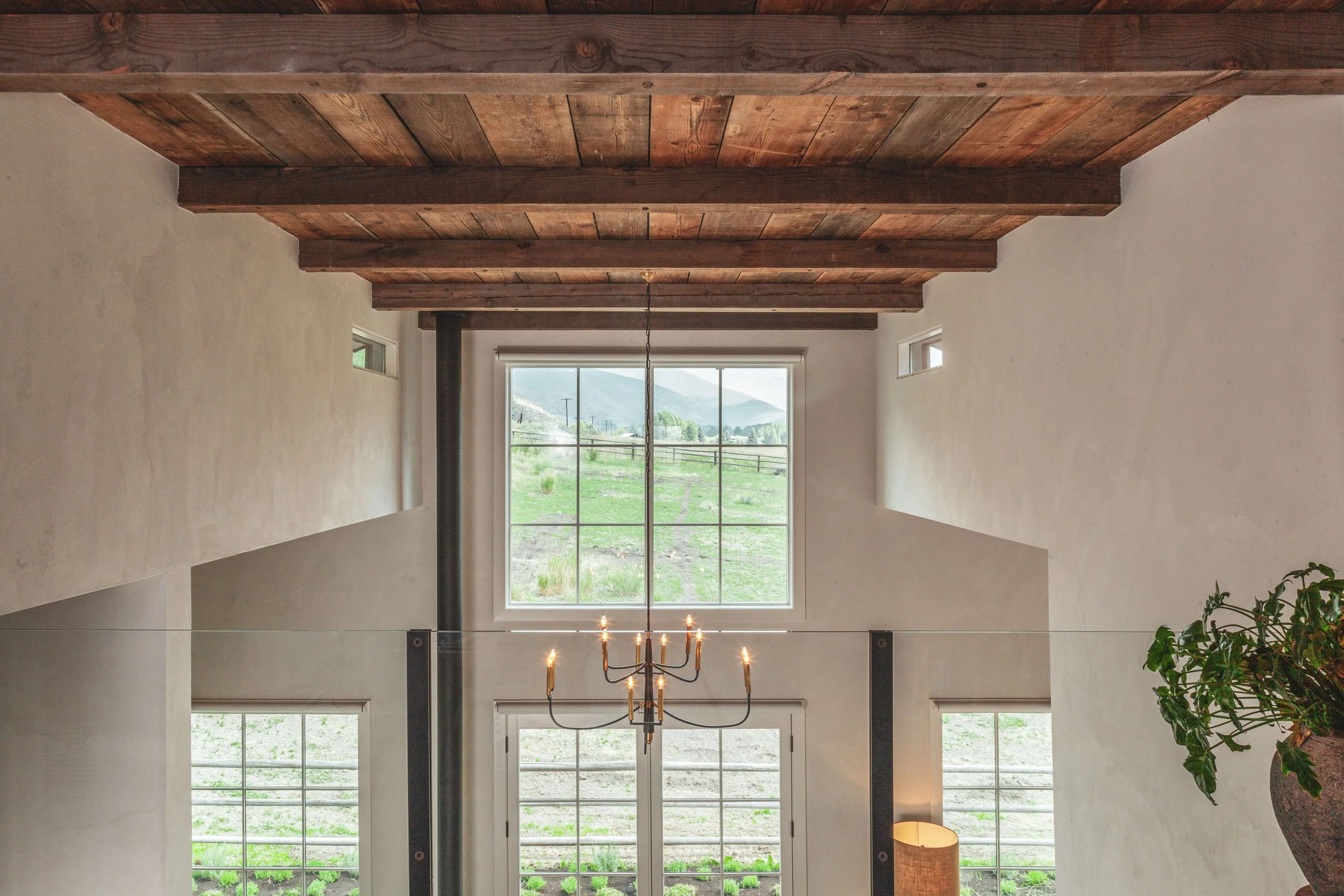 Interior view of a room with a wooden ceiling and large window showing a landscape of hills and greenery outside, a chandelier hanging from the ceiling, and a potted plant in the corner.