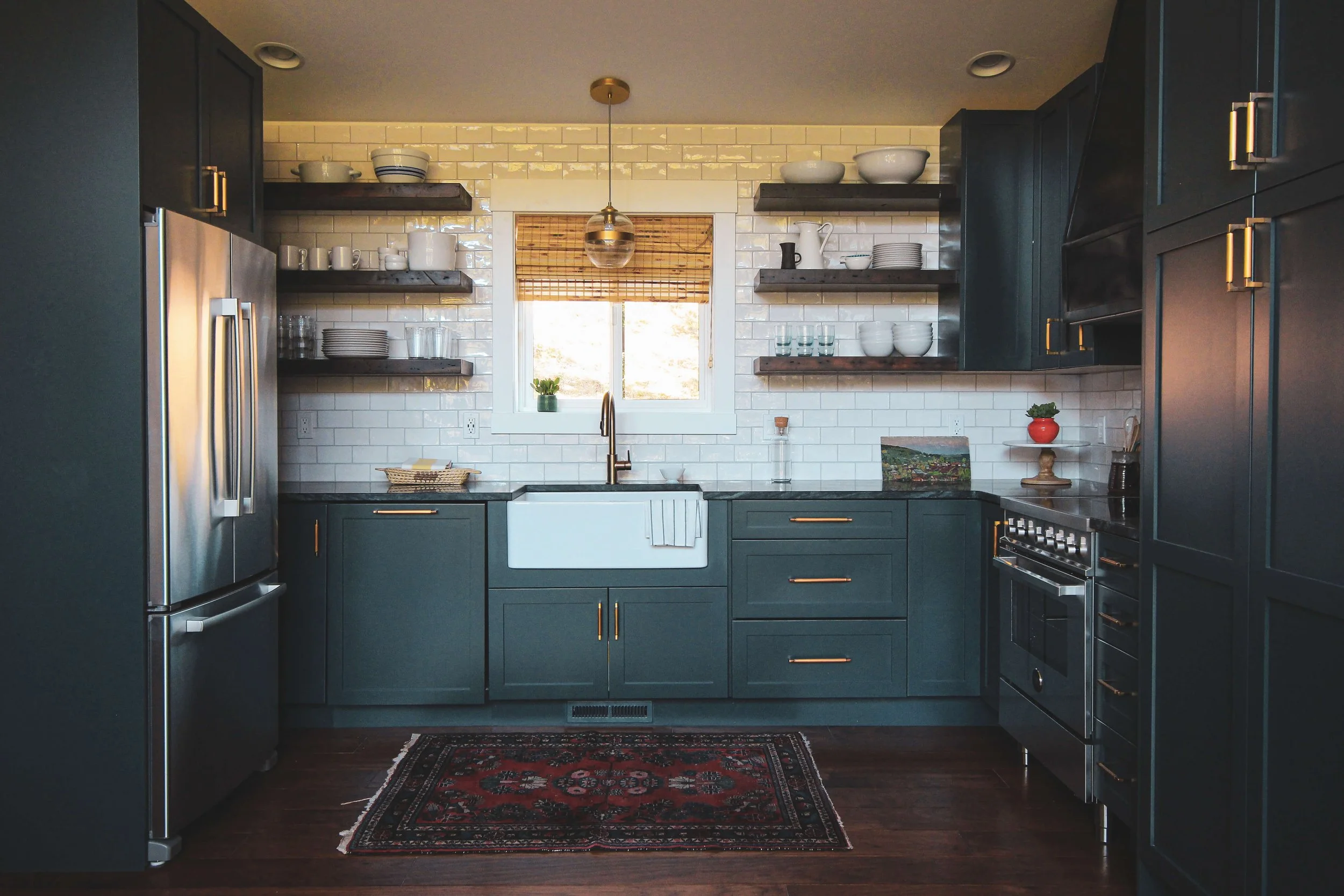 A modern kitchen with dark teal cabinets, open wooden shelves, white subway tile backsplash, stainless steel appliances, and a window with a bamboo shade. Decor includes bowls, cups, a plant, and small decorative items.