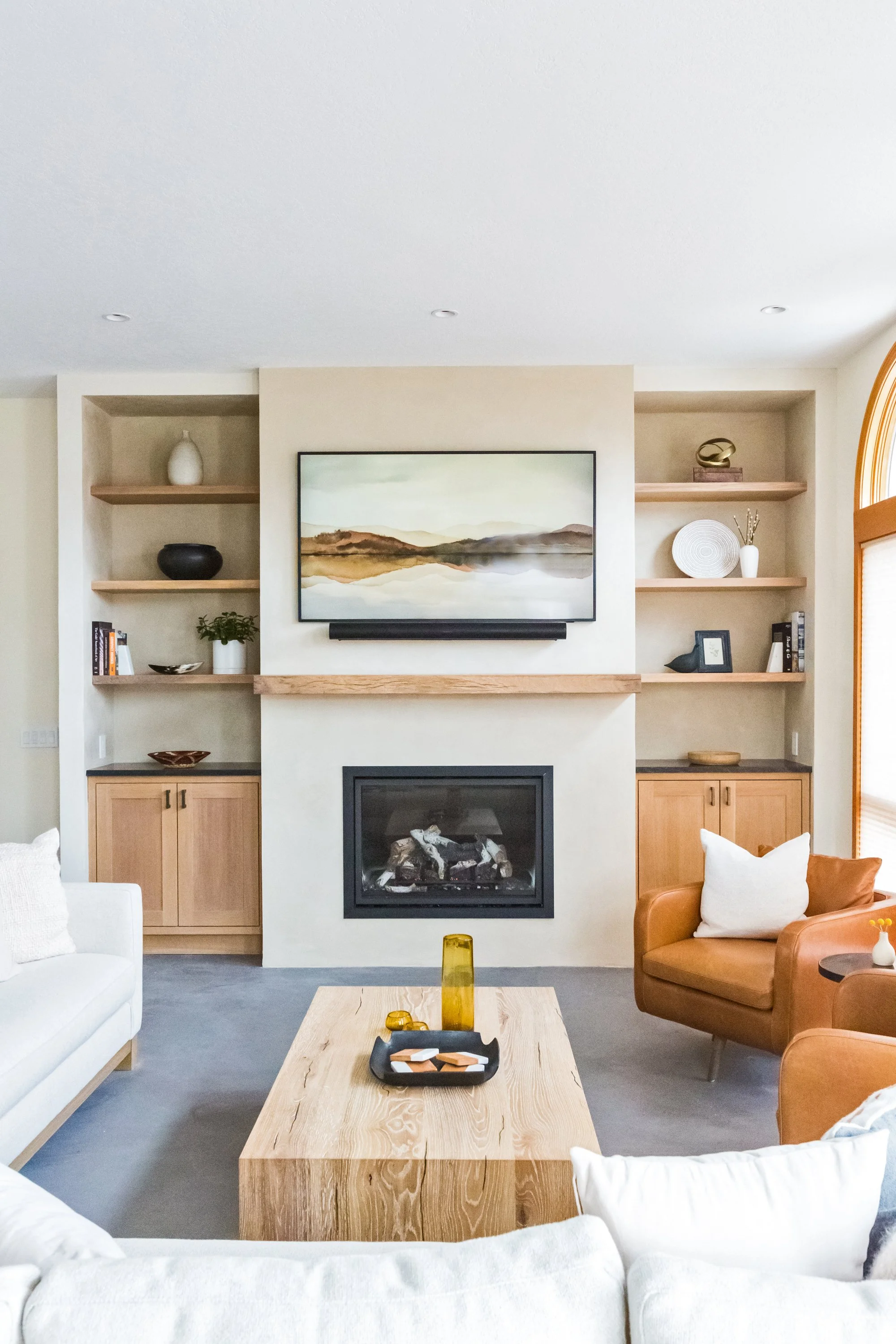 Living room with a fireplace, wall-mounted TV, built-in shelves with decorative items, and a seating area with white and brown armchairs around a wooden coffee table.