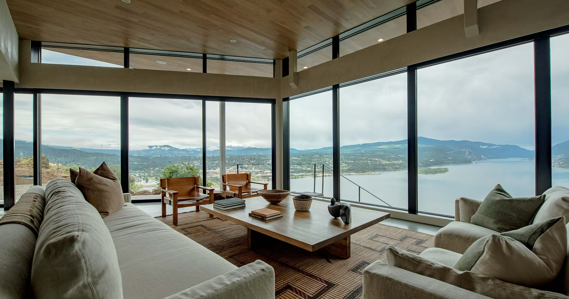 Living room with large floor-to-ceiling windows offering panoramic mountain and lake views, beige sofas, wooden chairs, and a coffee table with books and decorative objects.