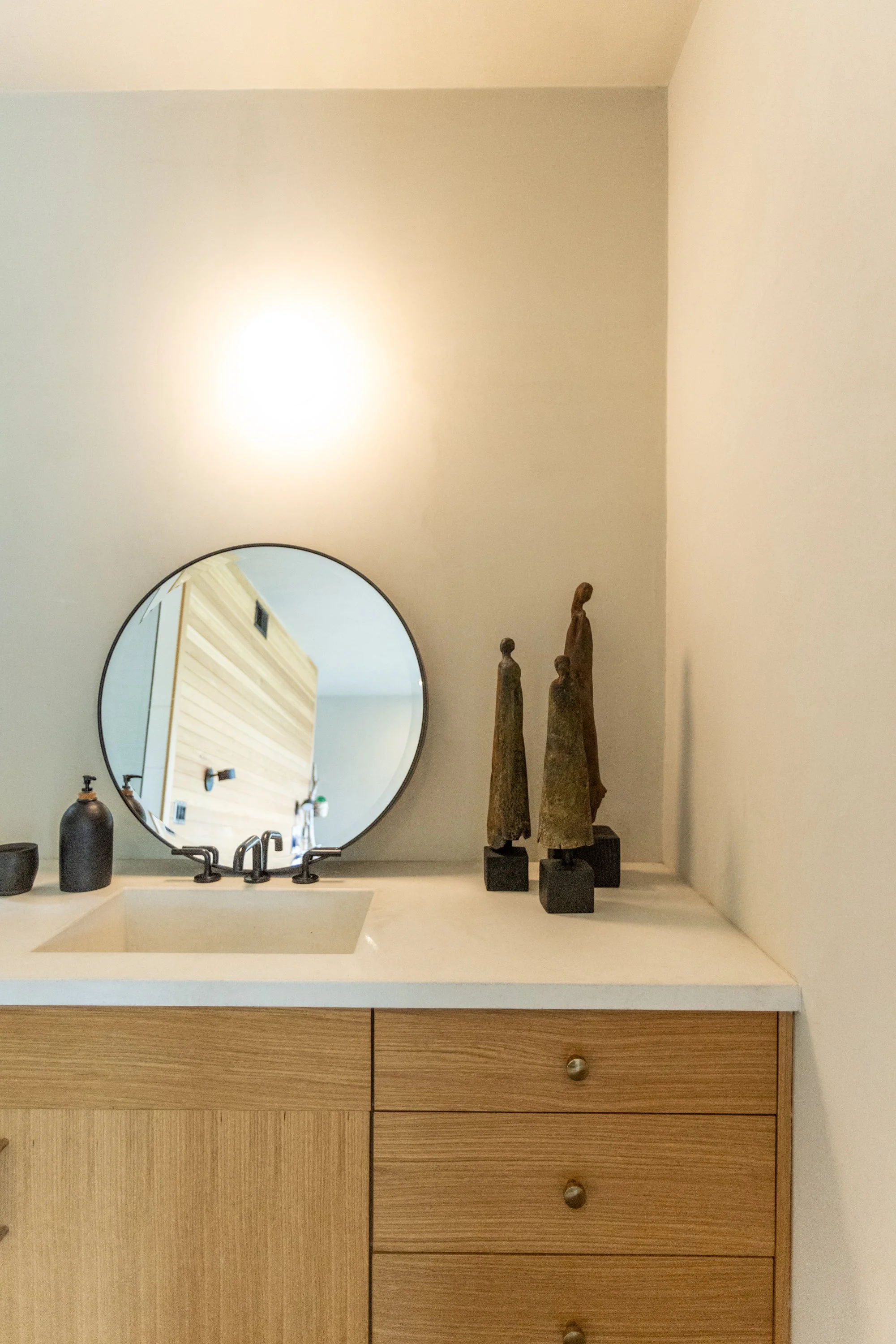 Bathroom vanity area with a round mirror, a tan countertop with a sink, black soap dispenser, and three tall decorative wooden statues on the right. Wooden drawers with round knobs below.