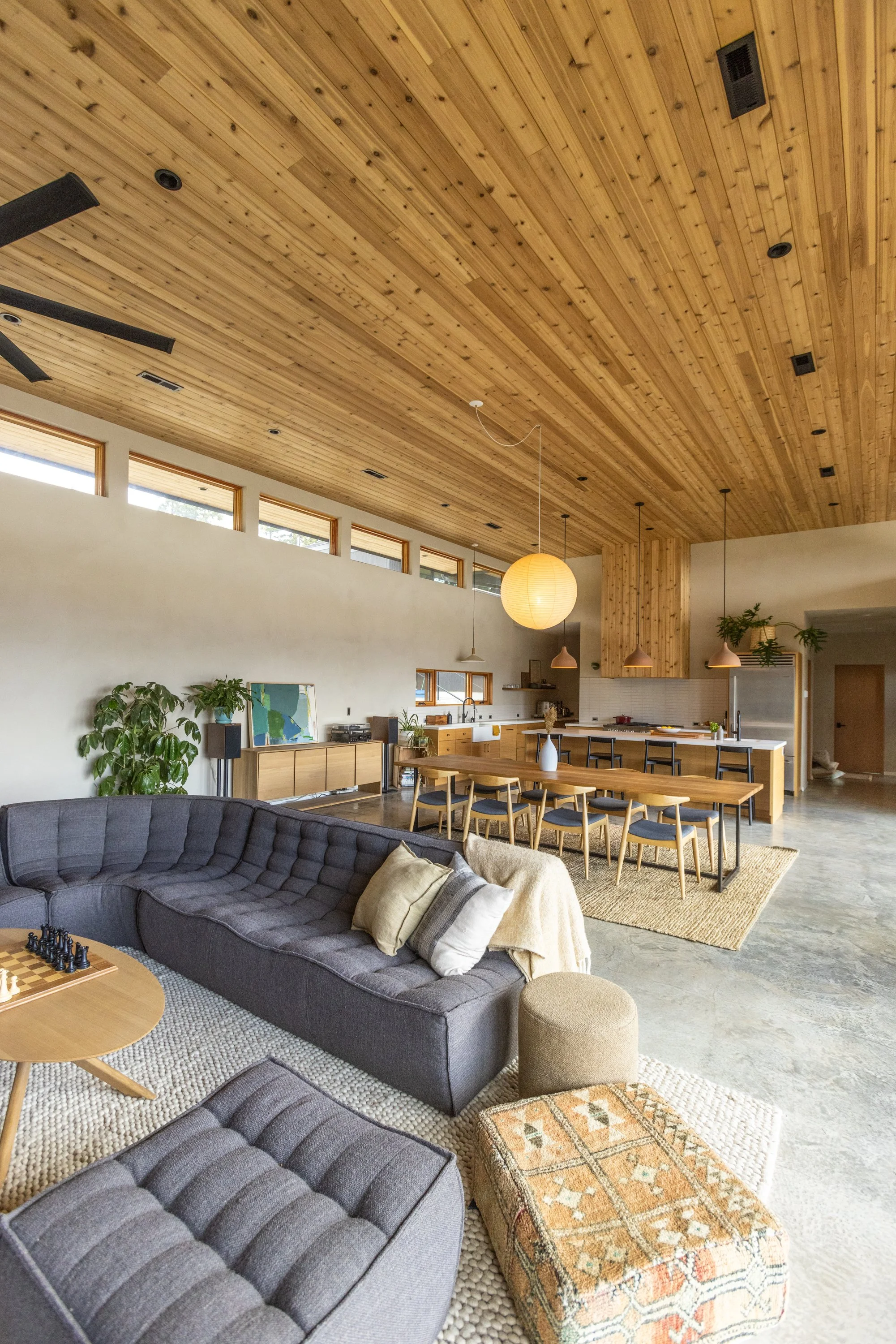 Open-concept living room and kitchen with wooden ceiling, large sectional sofa, patterned ottoman, dining table, pendant lights, and indoor plants.