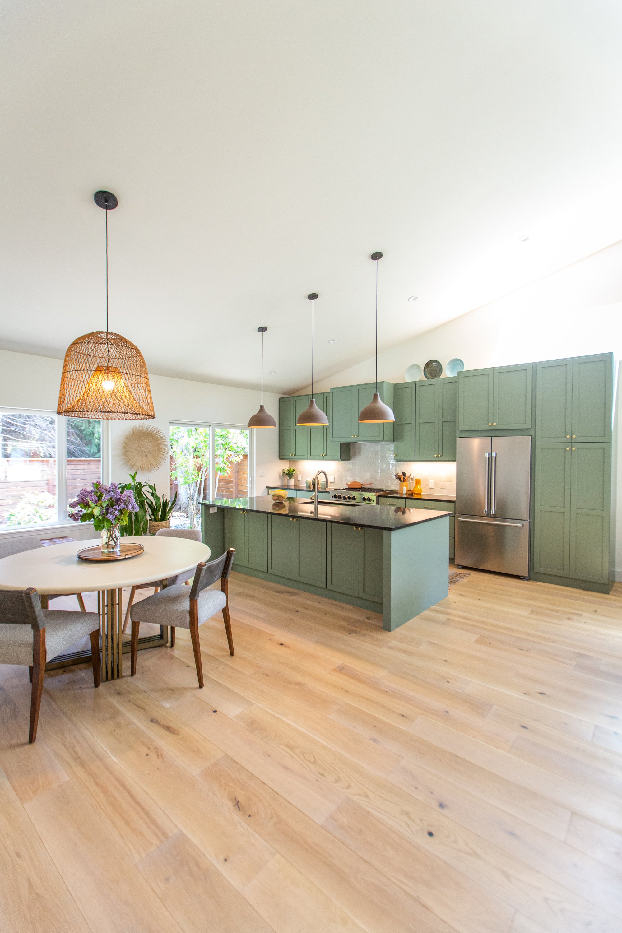 Kitchen with green cabinets, stainless steel refrigerator, white marble island, wooden floor, three hanging pendant lights, window with garden view, round dining table with chairs, flowers in a vase, and decorative plates on top of the cabinets.