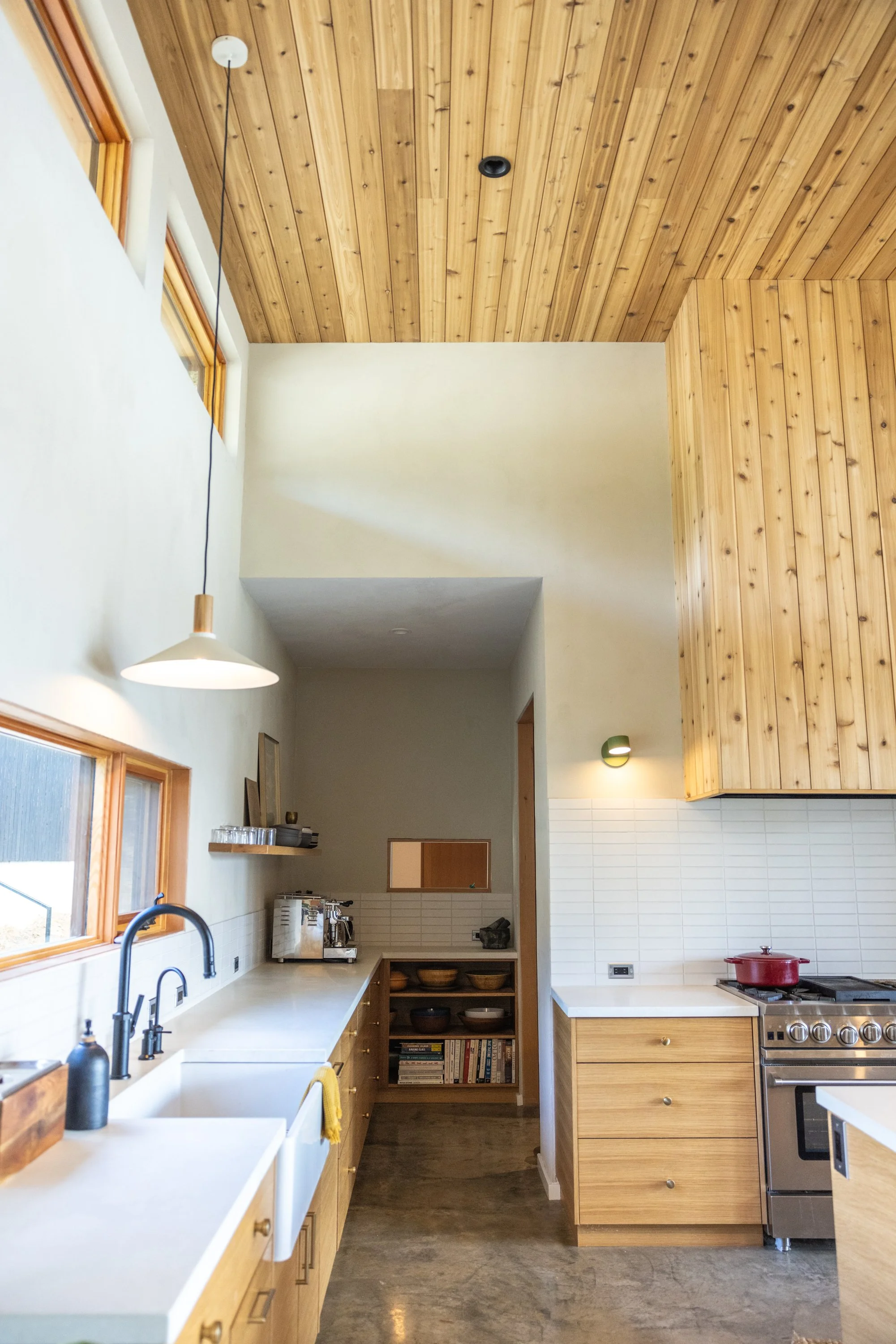 A modern kitchen with wooden cabinetry and a white countertop, featuring a sink, a coffee maker, and open shelves with dishes. There is a window with natural light, hanging pendant light, and a stove with a red pot. The ceiling has wooden paneling.