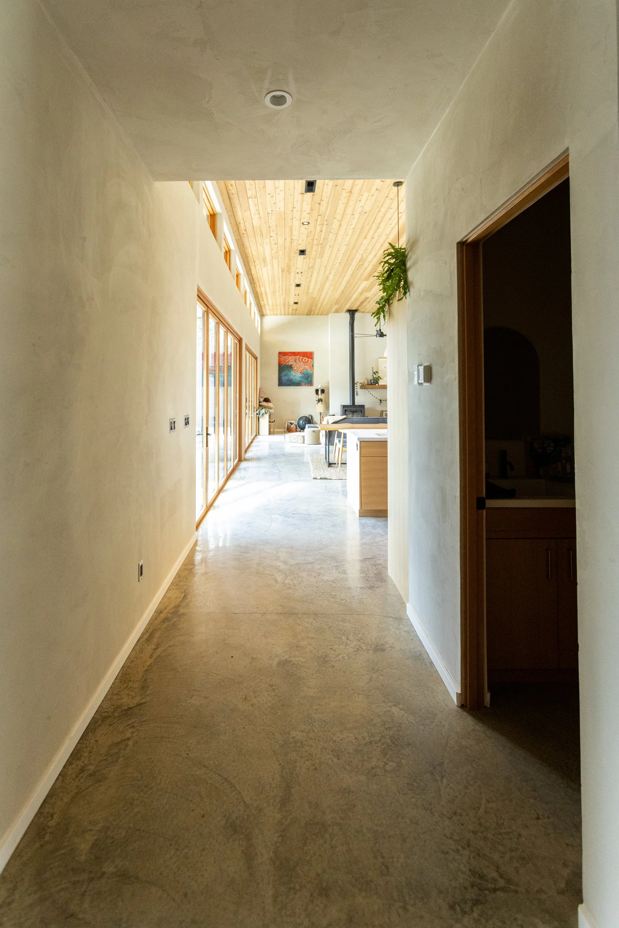 Empty hallway with concrete floor, cream-colored walls, and wooden ceiling. Natural light coming through glass doors on the left, with artwork and plants visible at the far end.