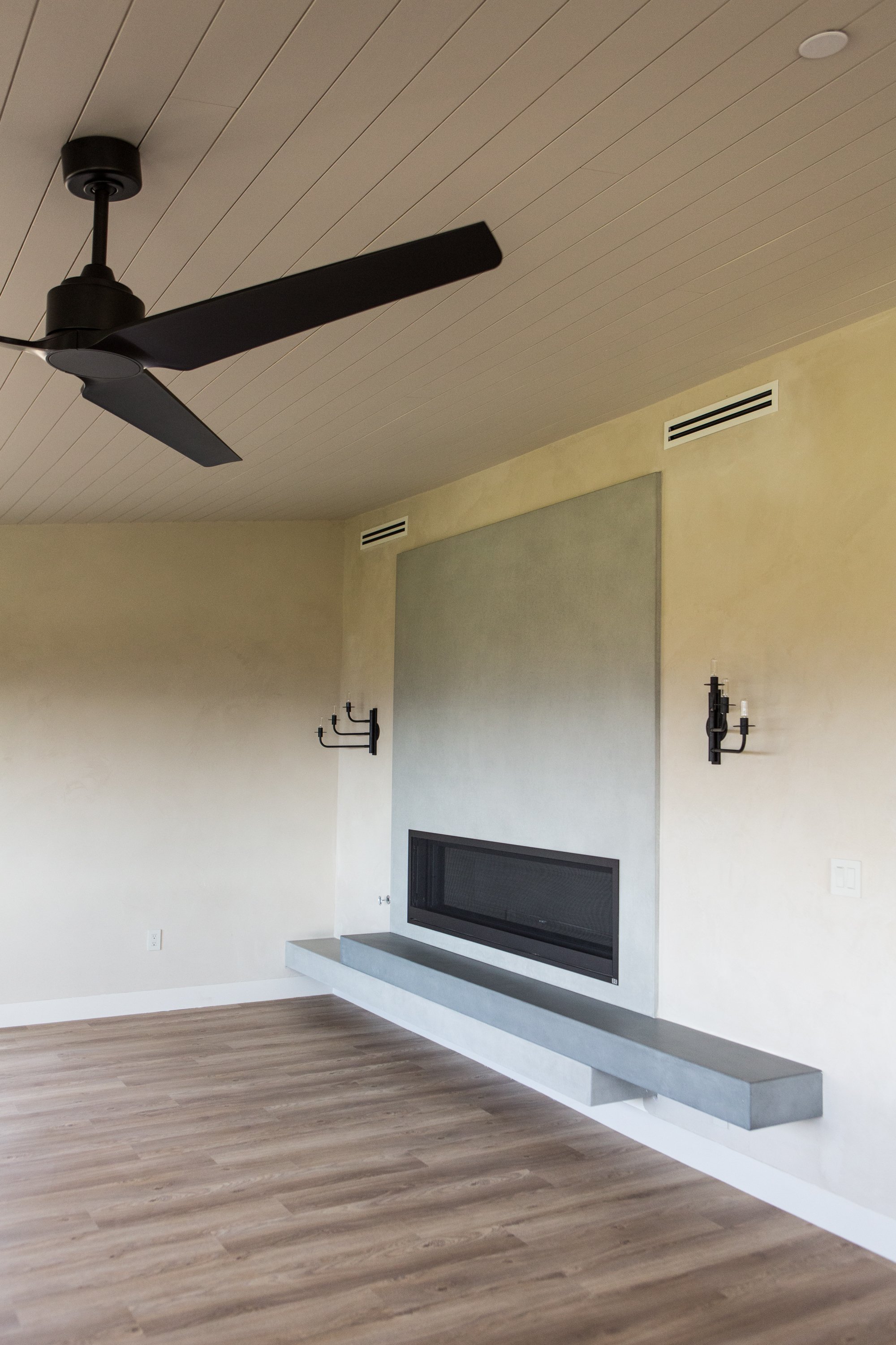 Modern living room with a ceiling fan, a fireplace, light-colored walls, and wooden flooring.