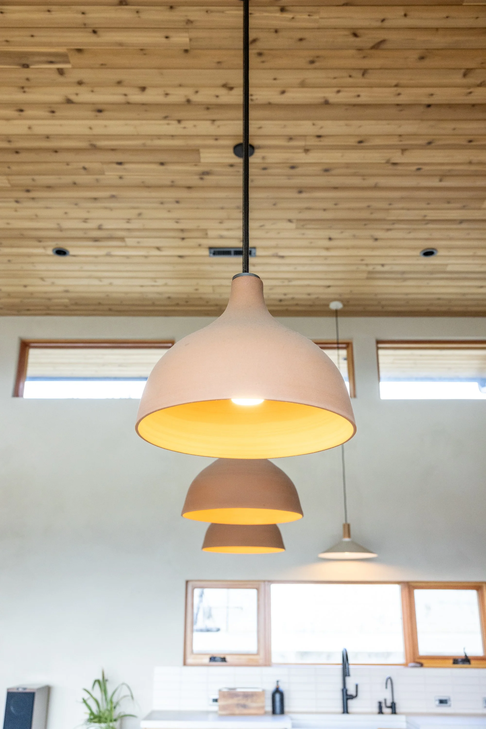 Multiple pendant lights hanging from a wooden ceiling over a modern kitchen with large windows, white countertops, black faucet, and backsplash.