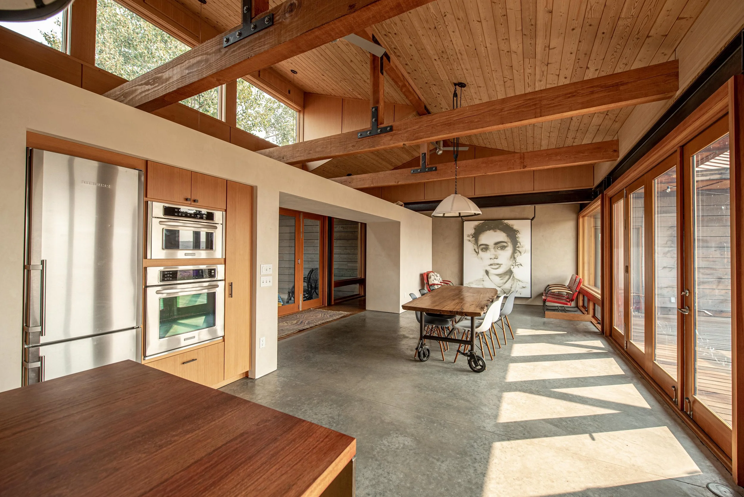 Modern kitchen and dining area with wooden ceiling beams, large windows, and a wooden dining table with white chairs. There is a large black-and-white portrait on the wall.