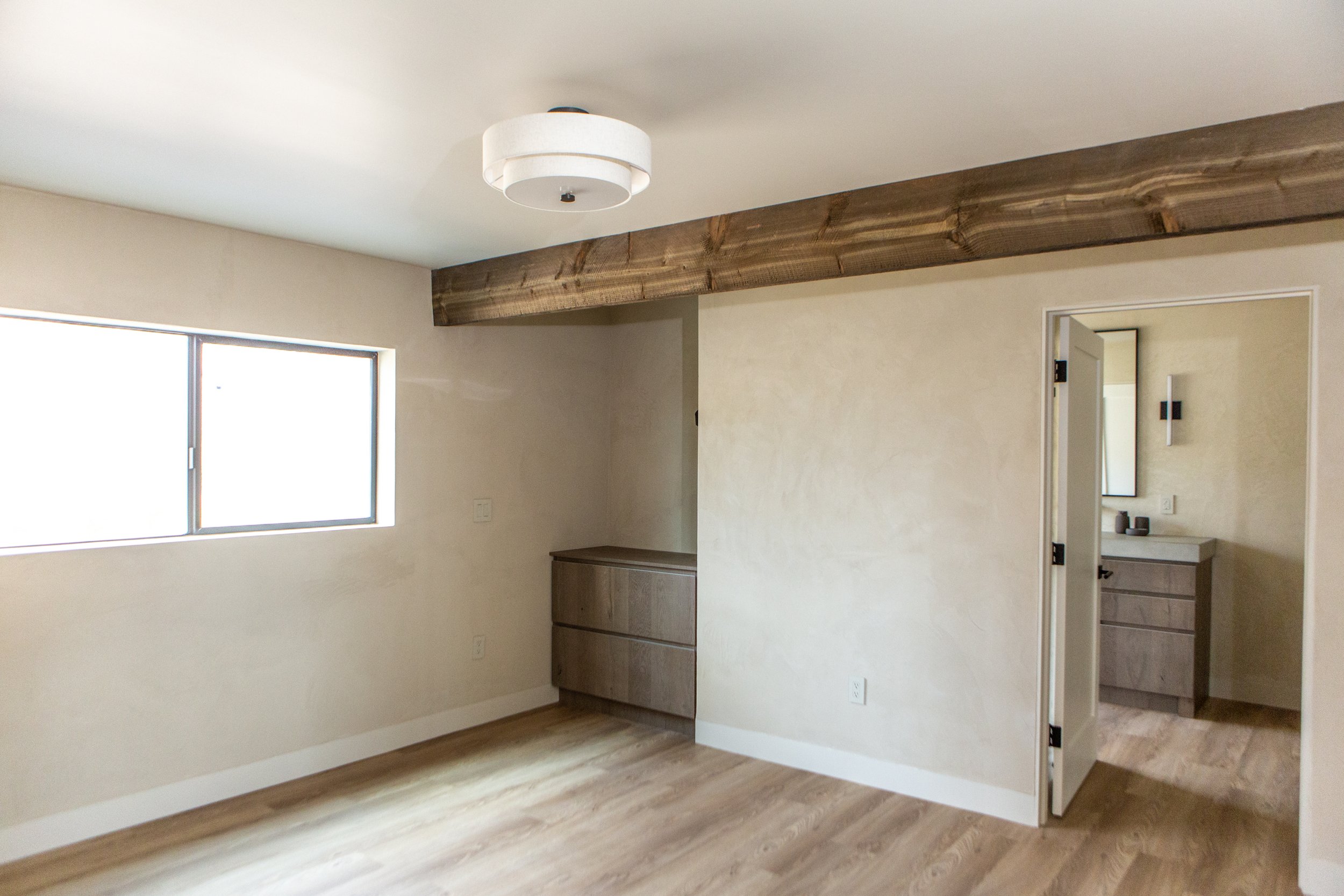 Empty room with wooden flooring, a window, a wooden beam on the ceiling, a small cabinet, and a door leading to a bathroom.