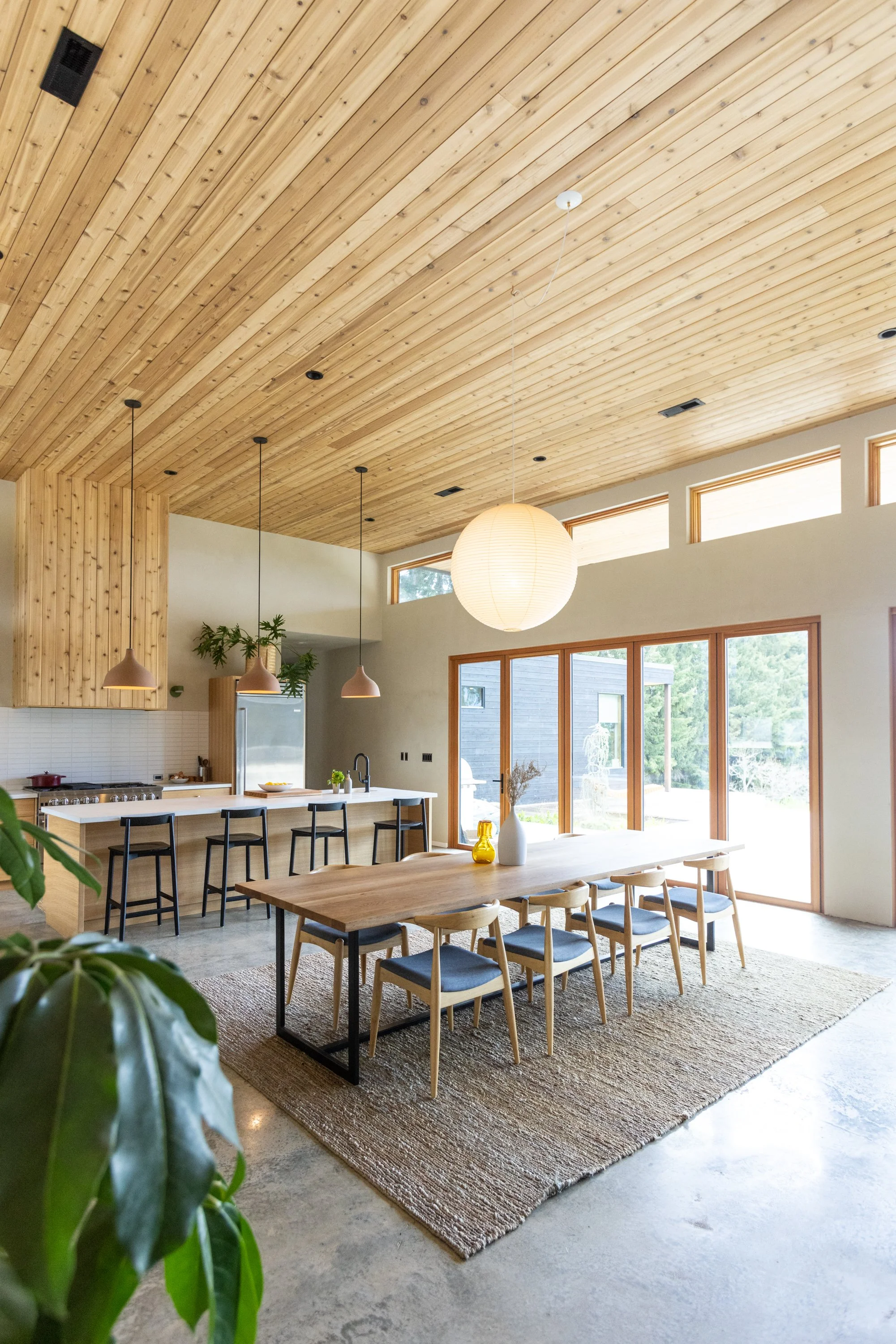 A spacious dining area with a large wooden table surrounded by wooden chairs. A textured rug is under the table. The room has a high wooden plank ceiling with large windows letting in natural light. There are pendant lights hanging above the kitchen 