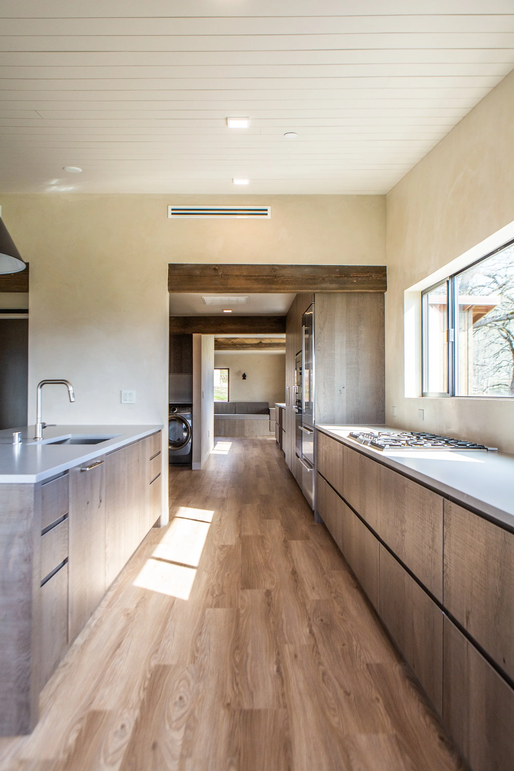 A modern kitchen with light wood cabinets, a white countertop with a sink, a large window, and wood flooring. There are recessed ceiling lights and visible through a doorway the living area with a gray sofa.