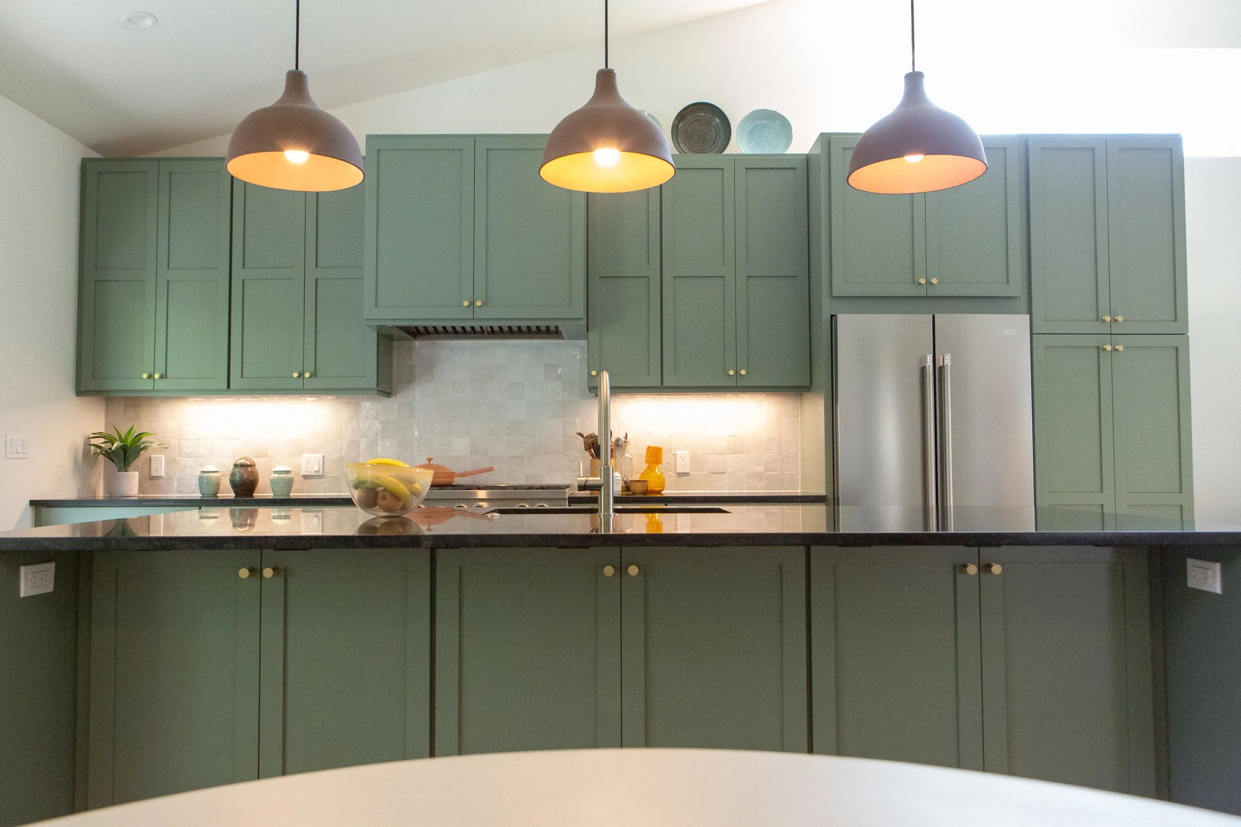 Kitchen with green cabinets, three black pendant lights, a fruit bowl, and decorative plates on top of the cabinets.