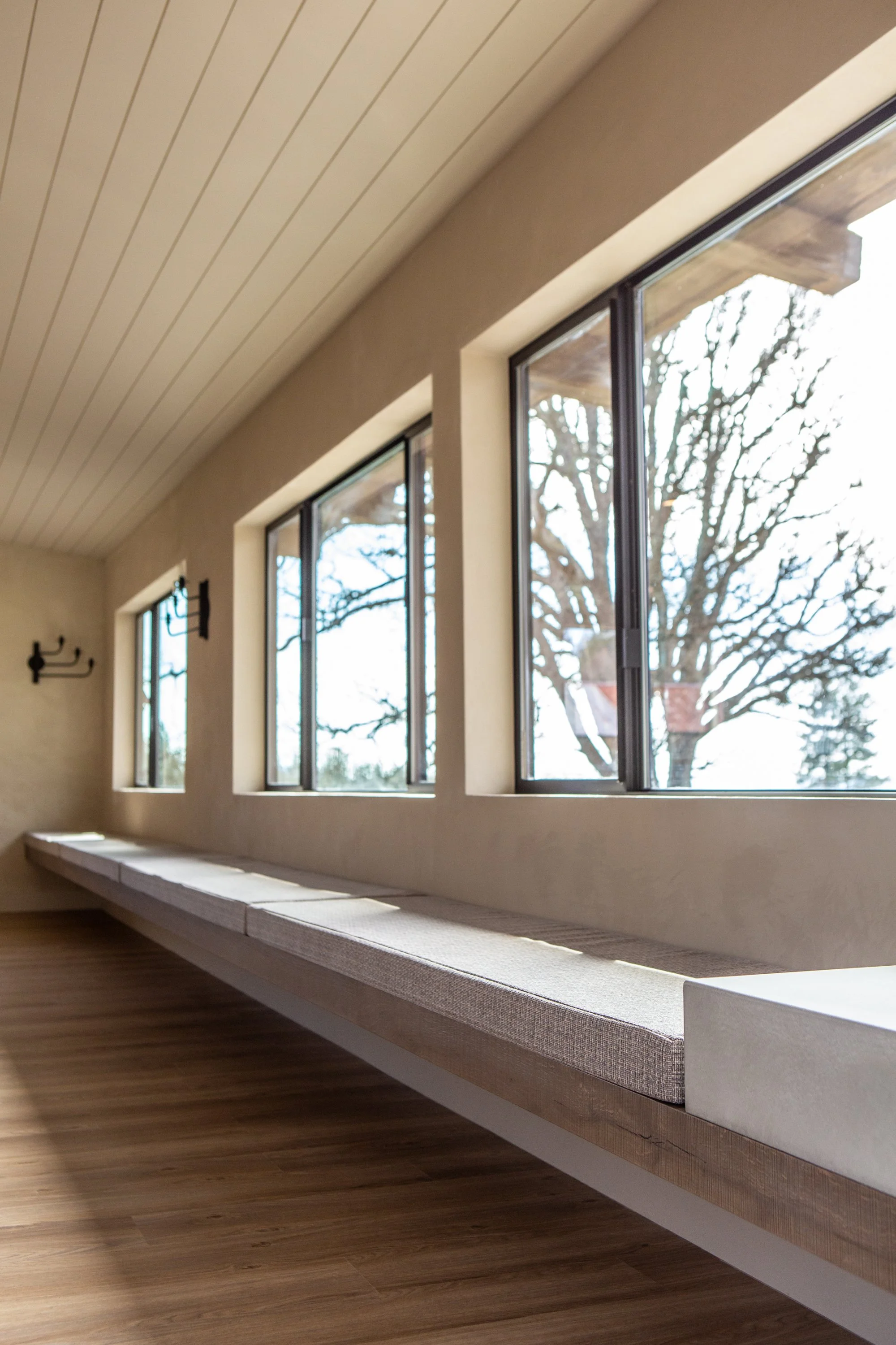 Interior view of a room with large windows looking out to a tree with bare branches, a built-in window seat with a cushioned surface, wood flooring, and a beige ceiling.
