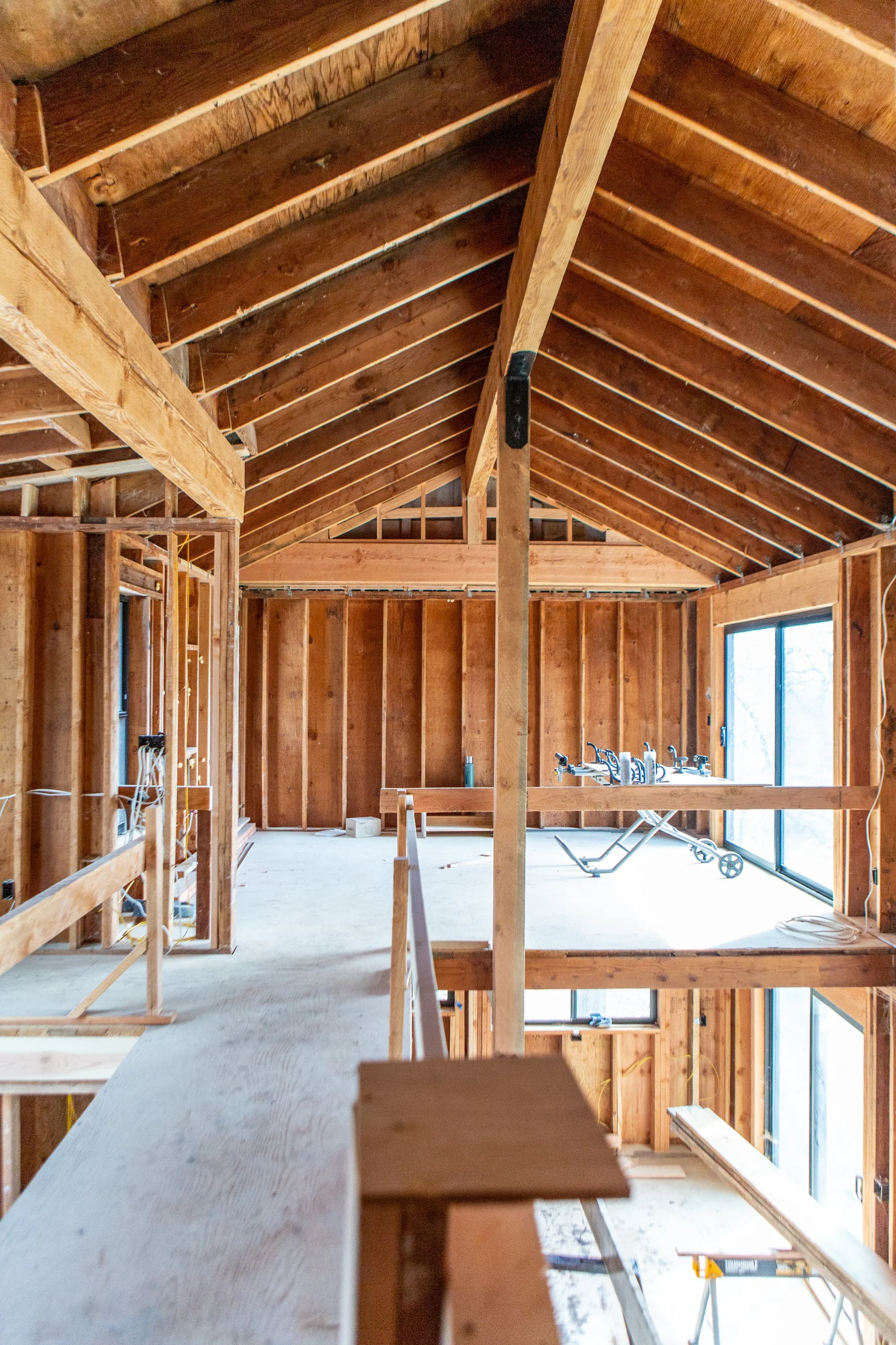 Interior of a house under construction, showing exposed wooden framing, partial wall and roof sheathing, and construction tools, with large windows letting in natural light.