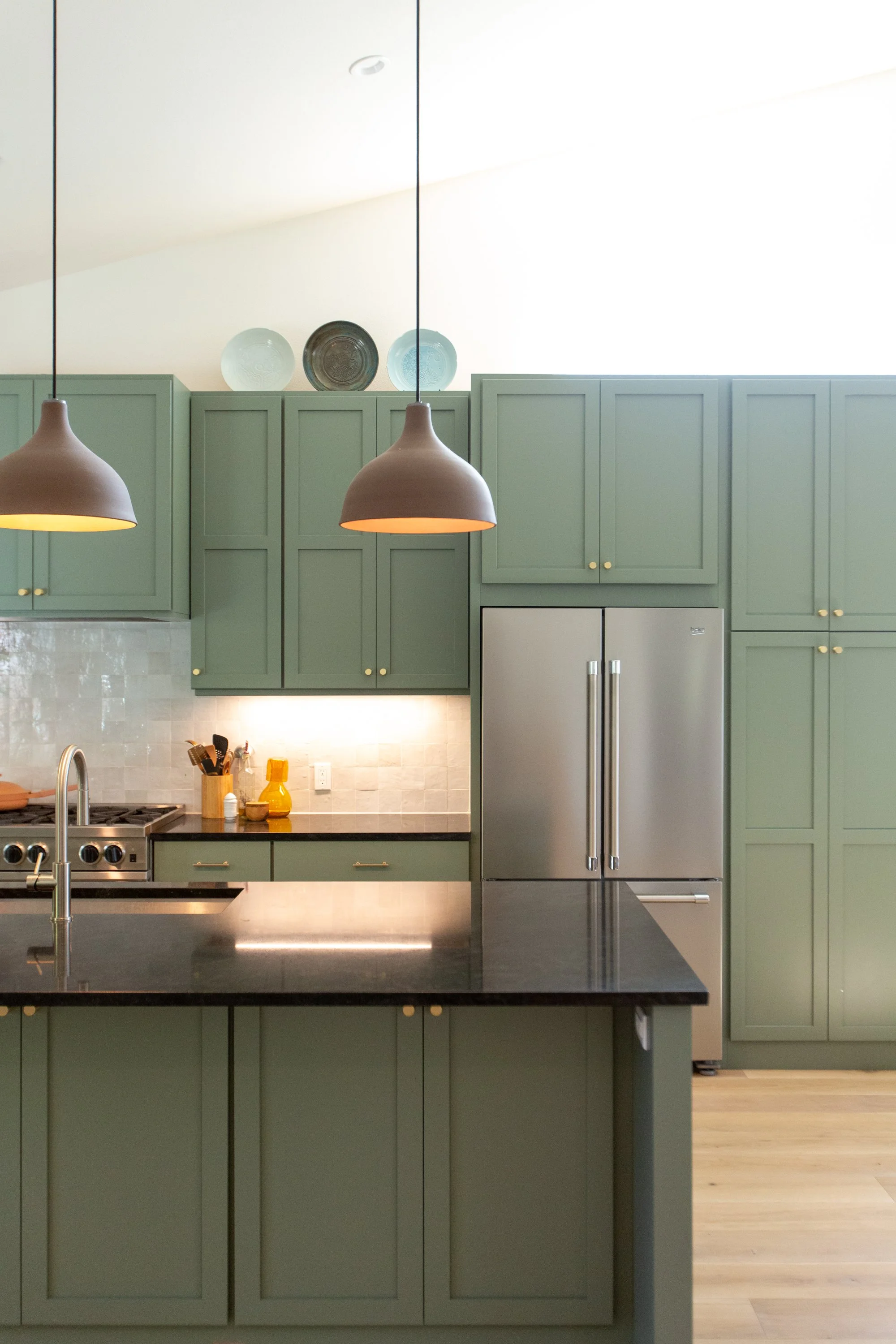 A modern kitchen with green cabinets, stainless steel refrigerator, black countertop, and light wood flooring. Two pendant lights hang above the counter, and decorative glass plates are displayed on top of the cabinets.