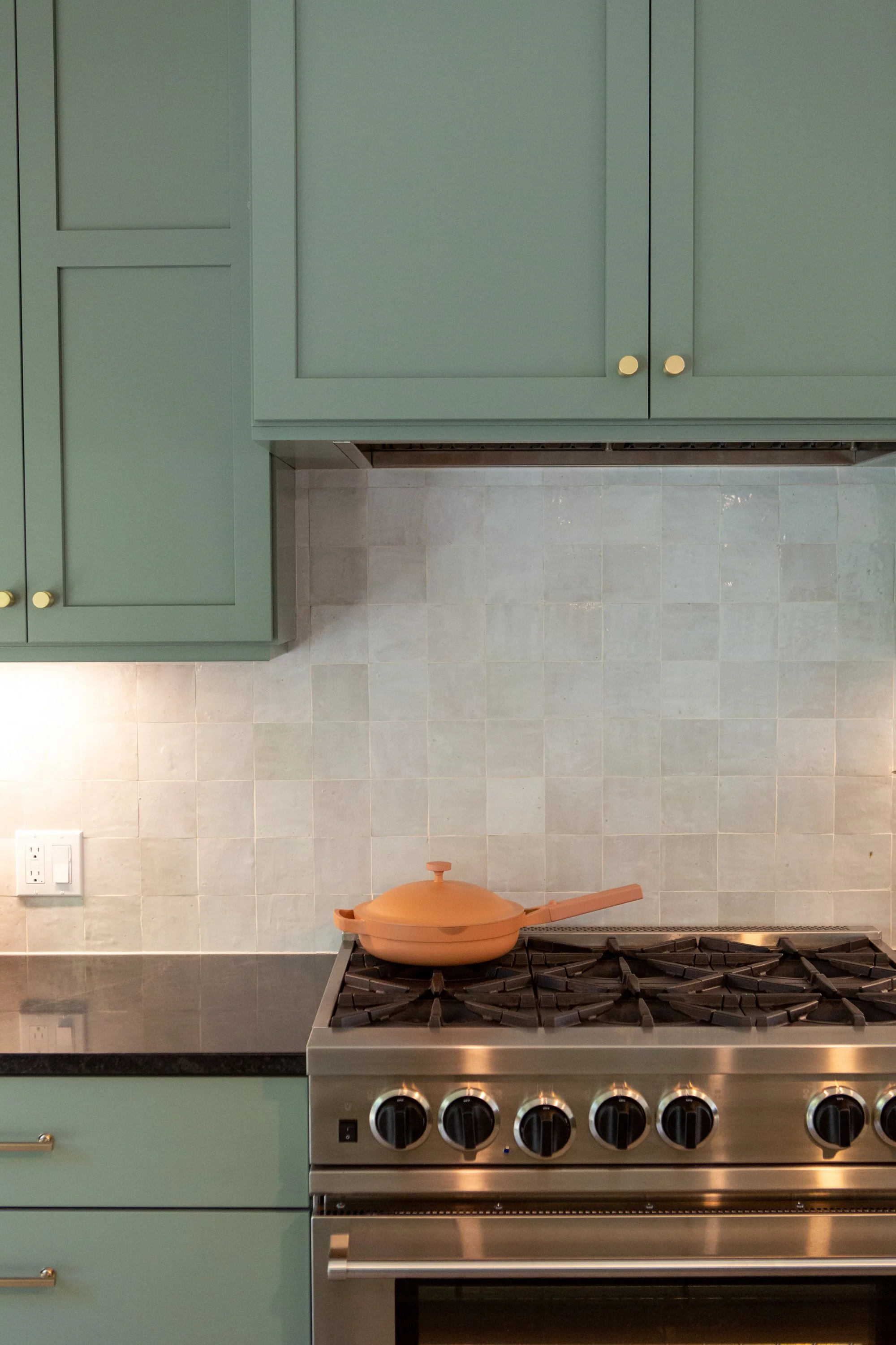 Green kitchen cabinets with gold knobs, a stove with black knobs, and a copper-colored frying pan on the stove. Light gray tiled wall in the background.
