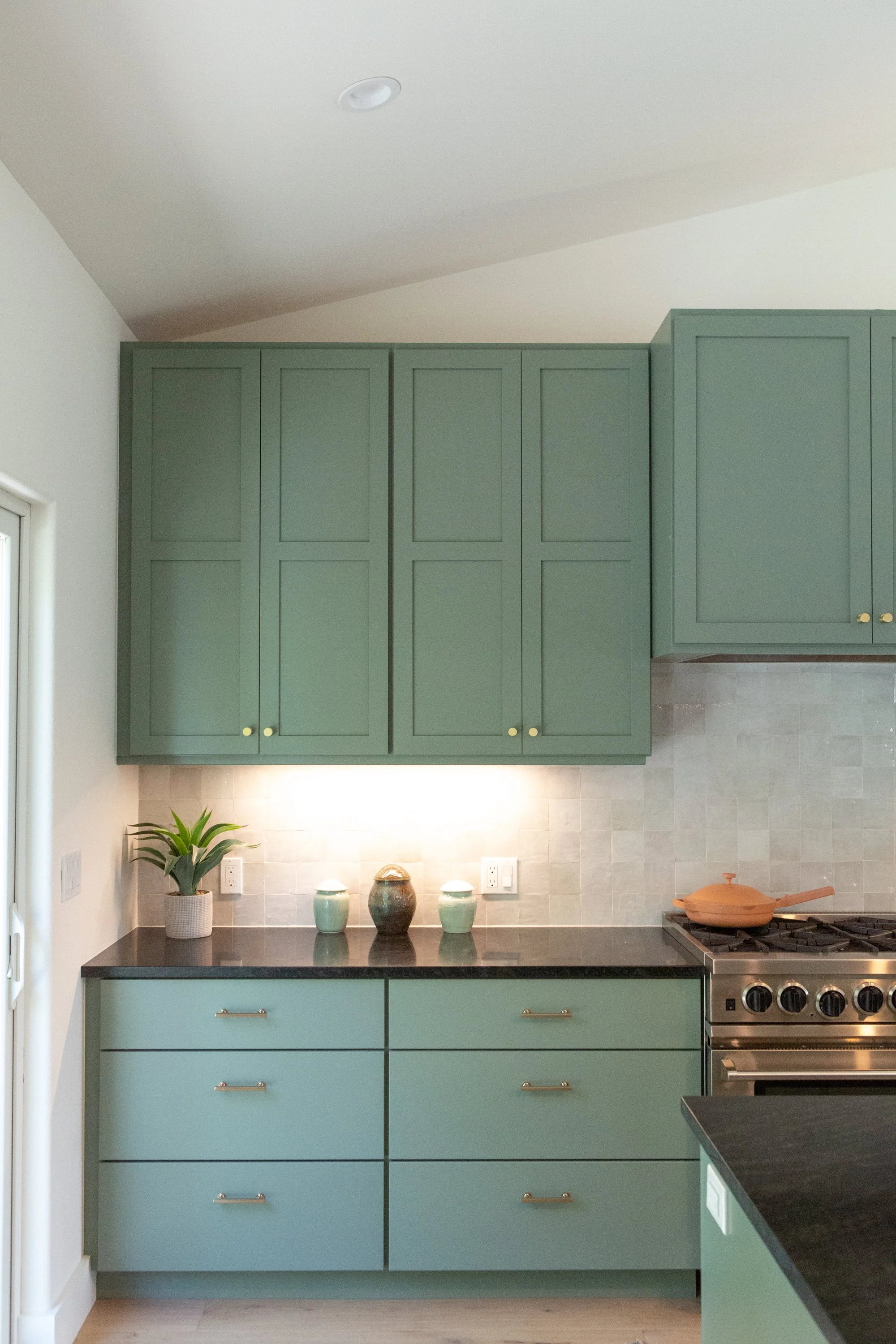 A modern kitchen with mint green cabinets, a black countertop, and a stove with a copper-colored skillet. Artwork and decorative vases sit on the counter, with a potted plant near a window on the left.