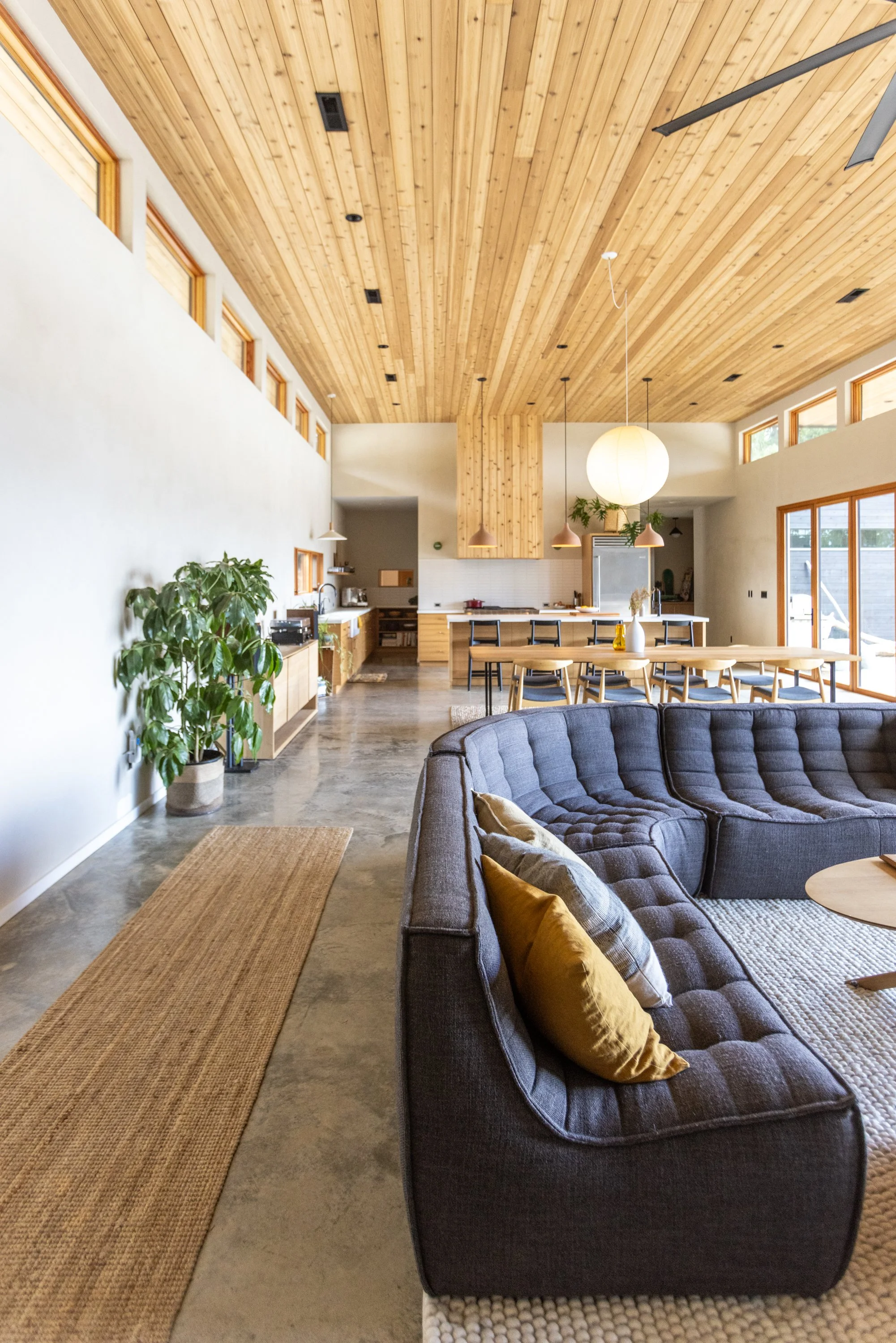 Open-concept living and dining area with high wooden ceiling, pendant lights, gray tufted sofa with gold pillows, large window, and indoor plants.