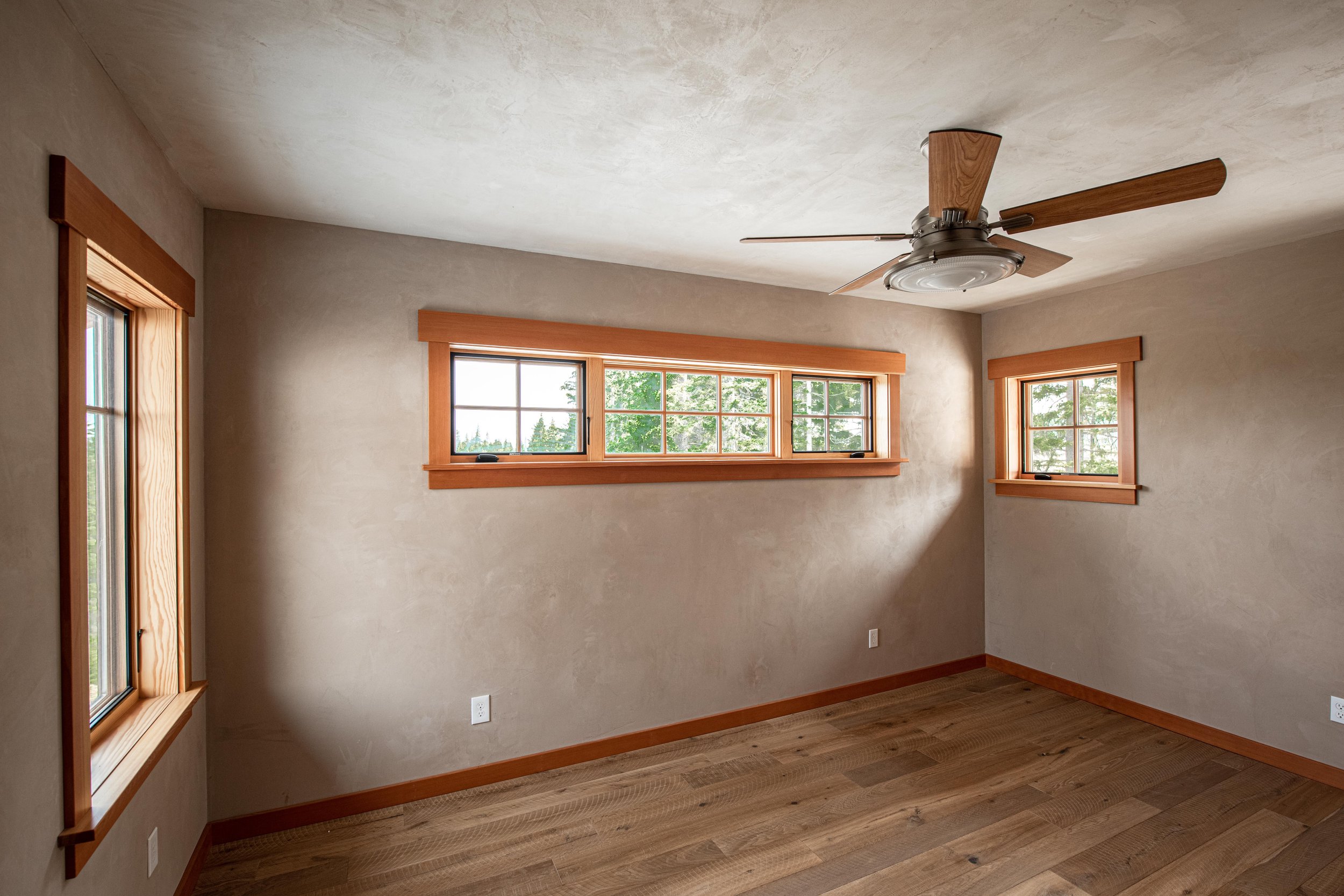 An empty room with beige walls, wooden window frames, sunlight coming through the windows, a ceiling fan with wooden blades, and hardwood flooring.