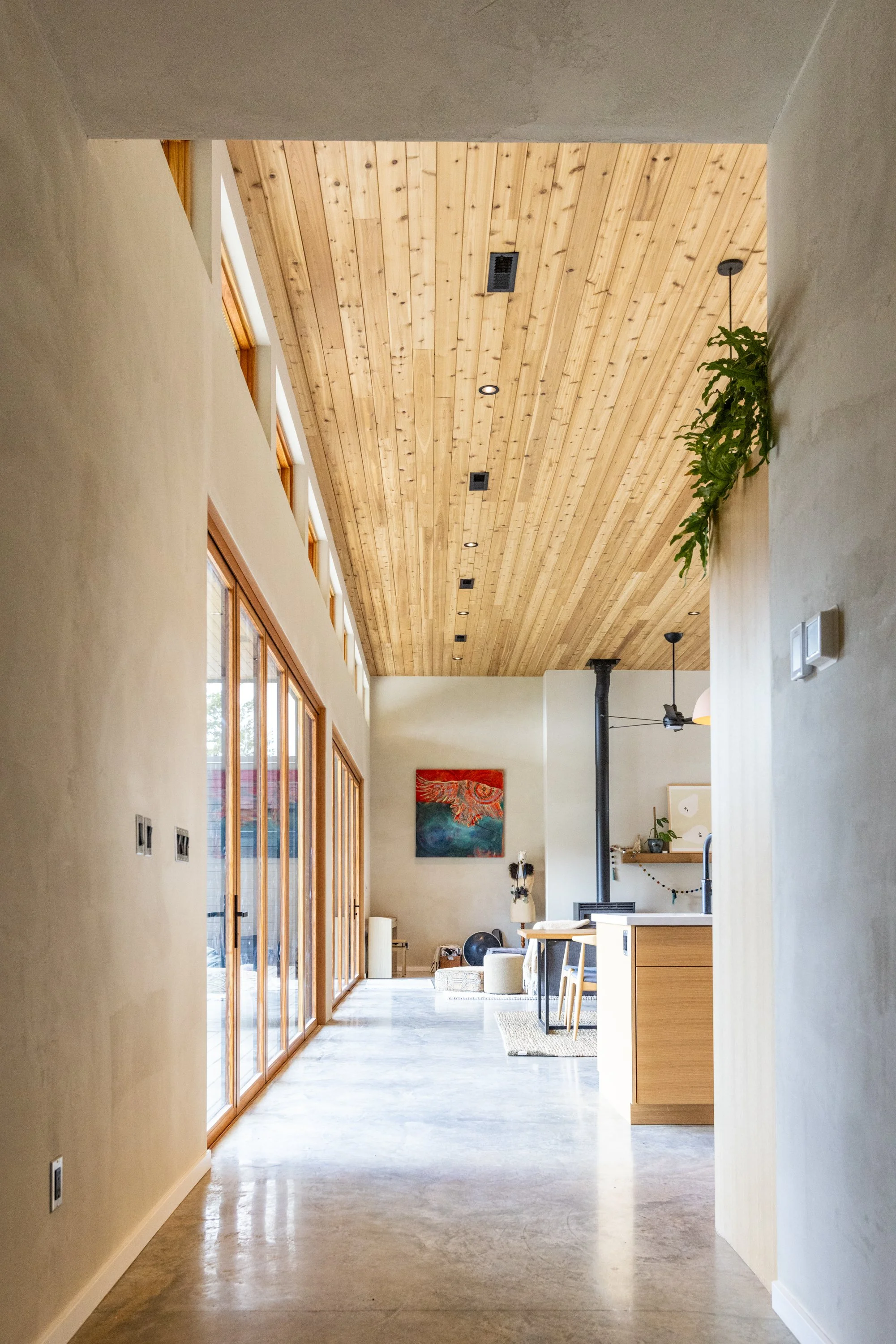Interior of a modern living space with a high wooden ceiling, large windows on the left, a cozy seating area with a painting on the wall, and a wood-burning stove in the background.