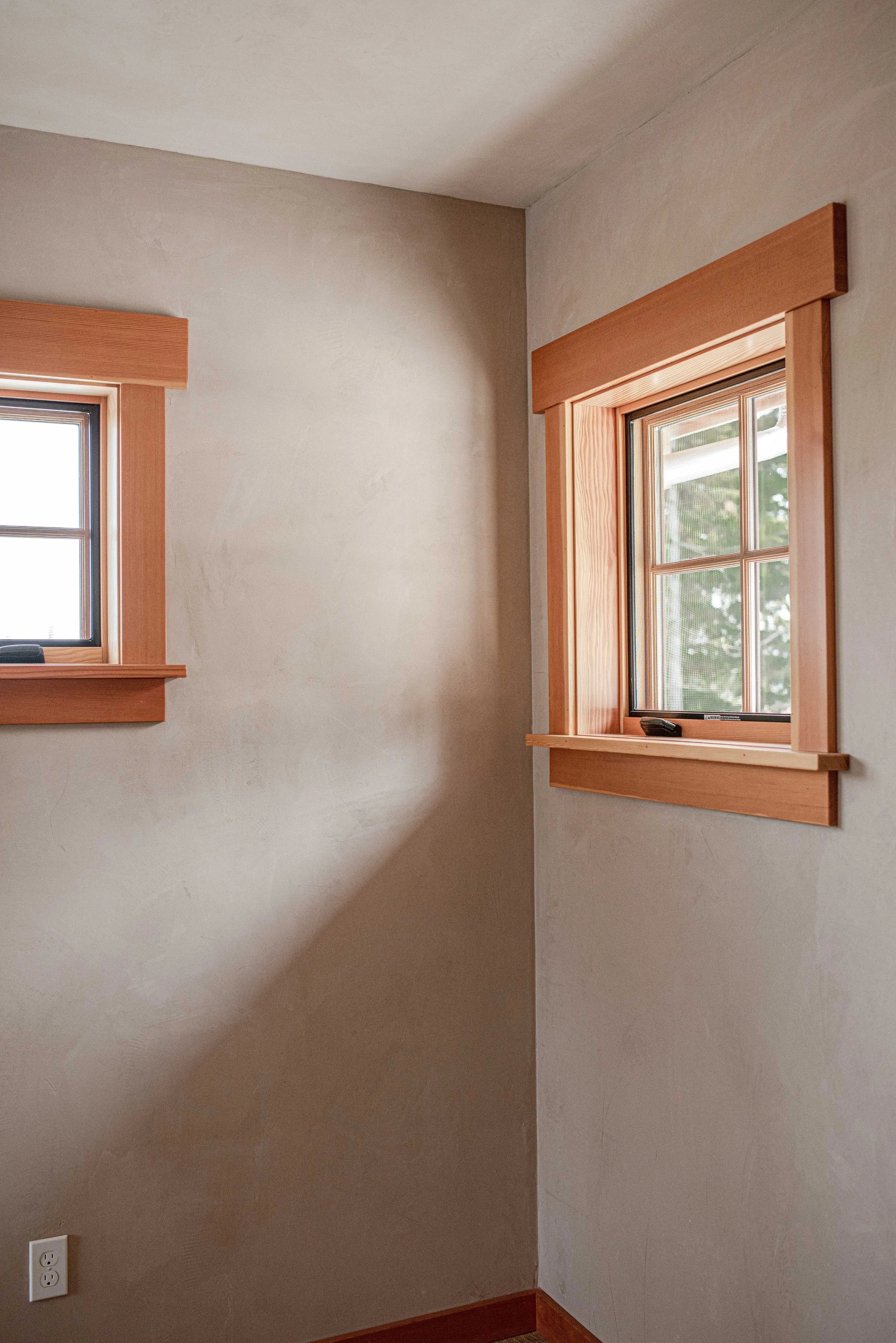 Two windows with wooden frames showing greenery outside, set in a beige wall with an electrical outlet at the bottom.