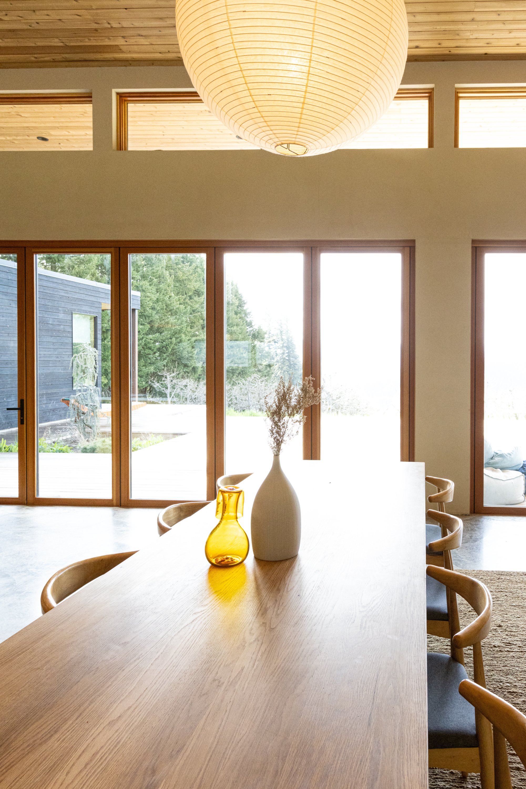 A modern dining room with a long wooden table, wooden chairs, large windows, and decorative vases on the table.