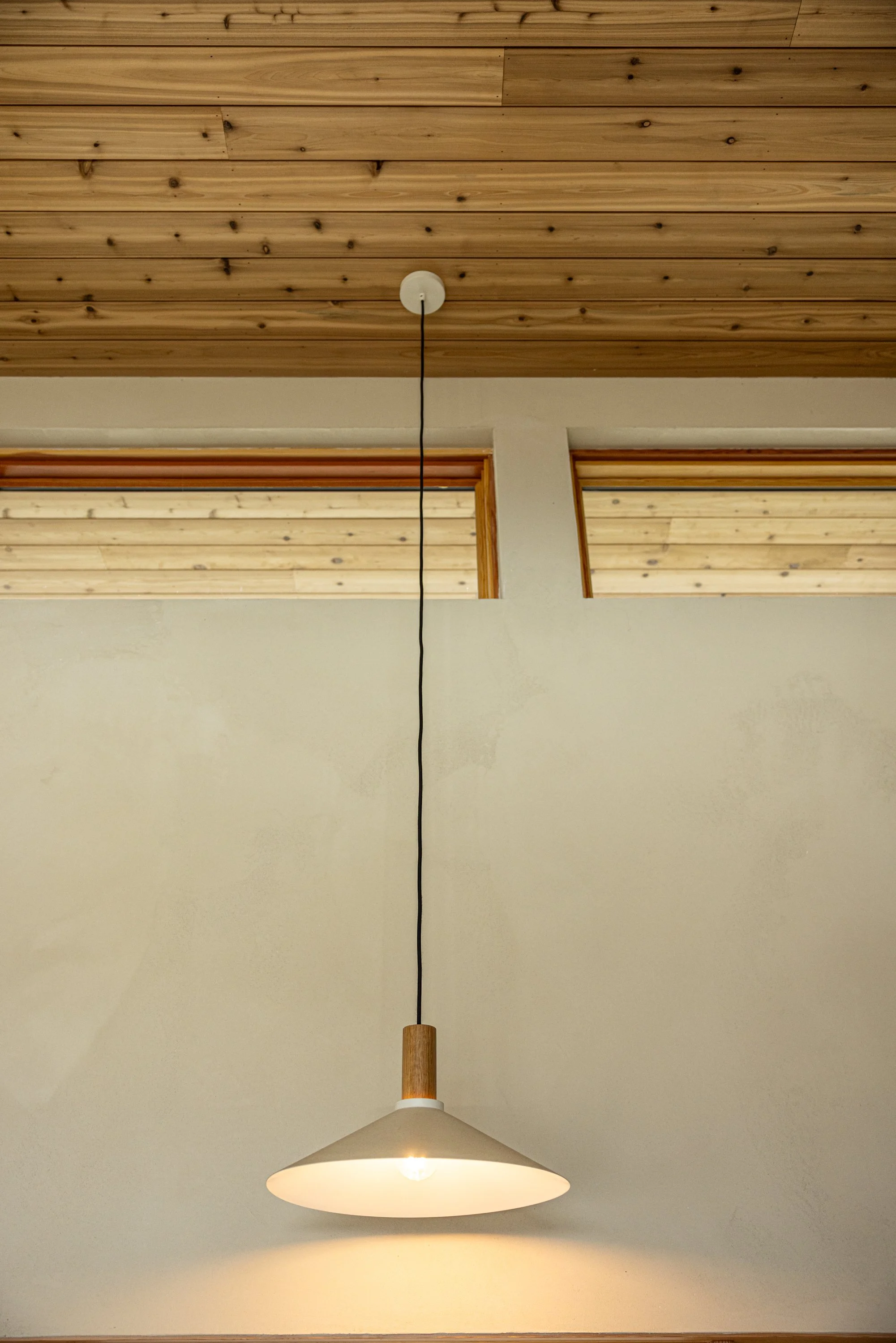 A white ceiling lamp hanging from a wooden ceiling with visible wood paneling. The lamp has a black cord and a wooden accent.
