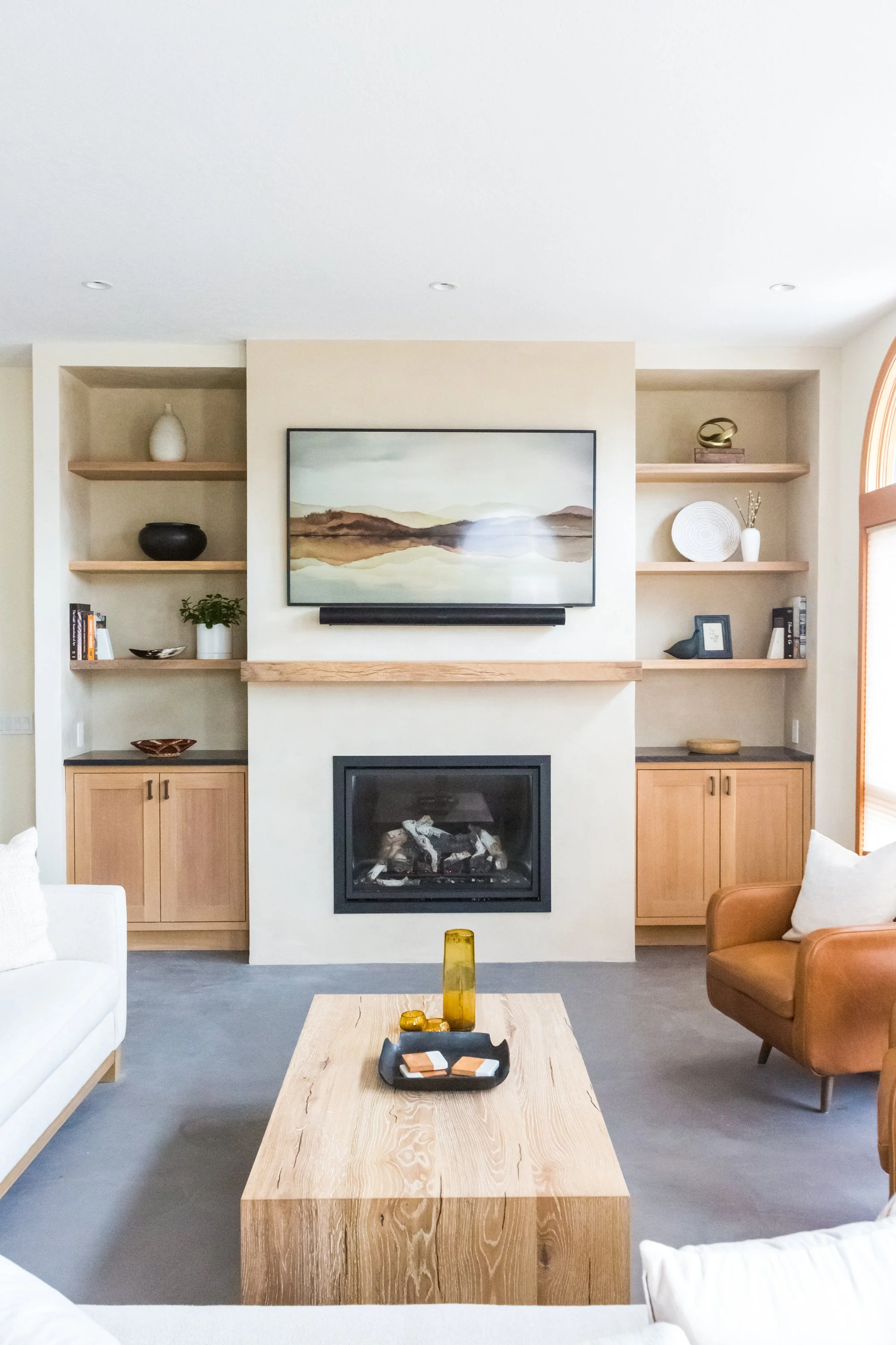 Living room with a fireplace, built-in shelves with decorative items, a wall-mounted television, a brown armchair, a white sofa, and a wooden coffee table with decorative objects.