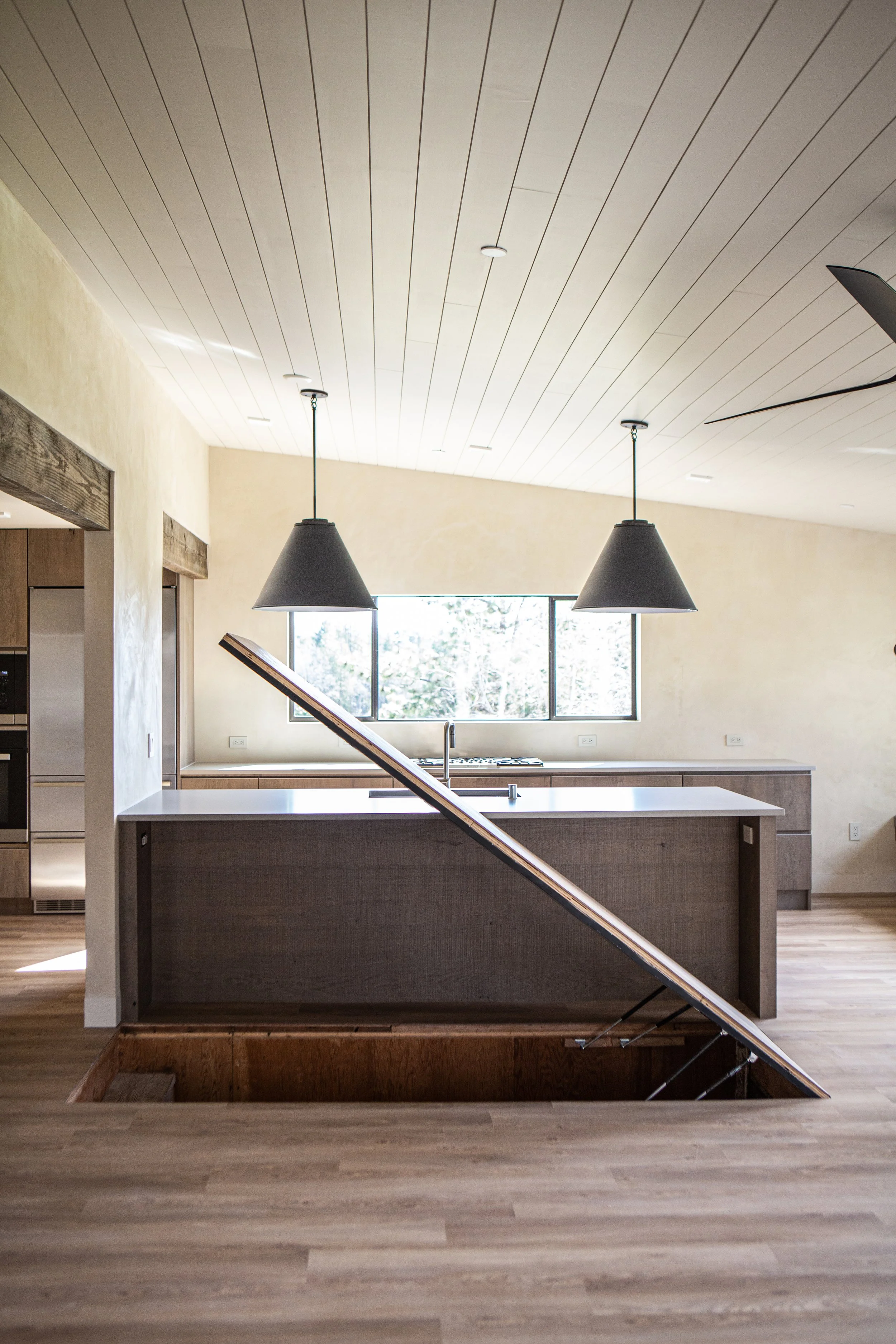 Interior of a modern kitchen with a hanging light fixture, a large window, a kitchen island with a sink, and an open staircase leading down.