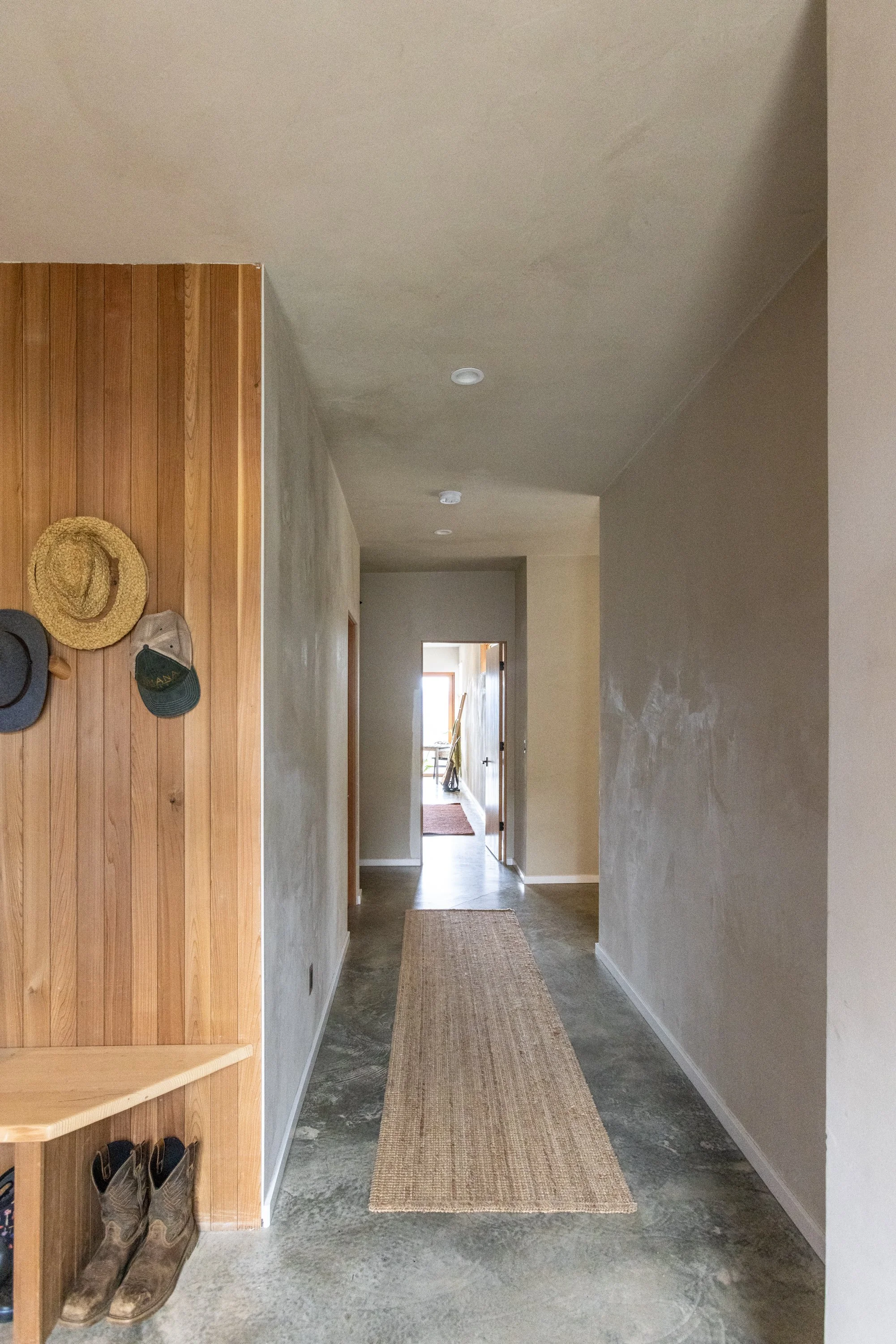 Hallway with beige textured walls, concrete floor, a brown woven rug, and ceiling-mounted lights. A doorway at the end leads to another room with natural light. To the left, a wooden wall section displays hats, including a straw hat and baseball caps