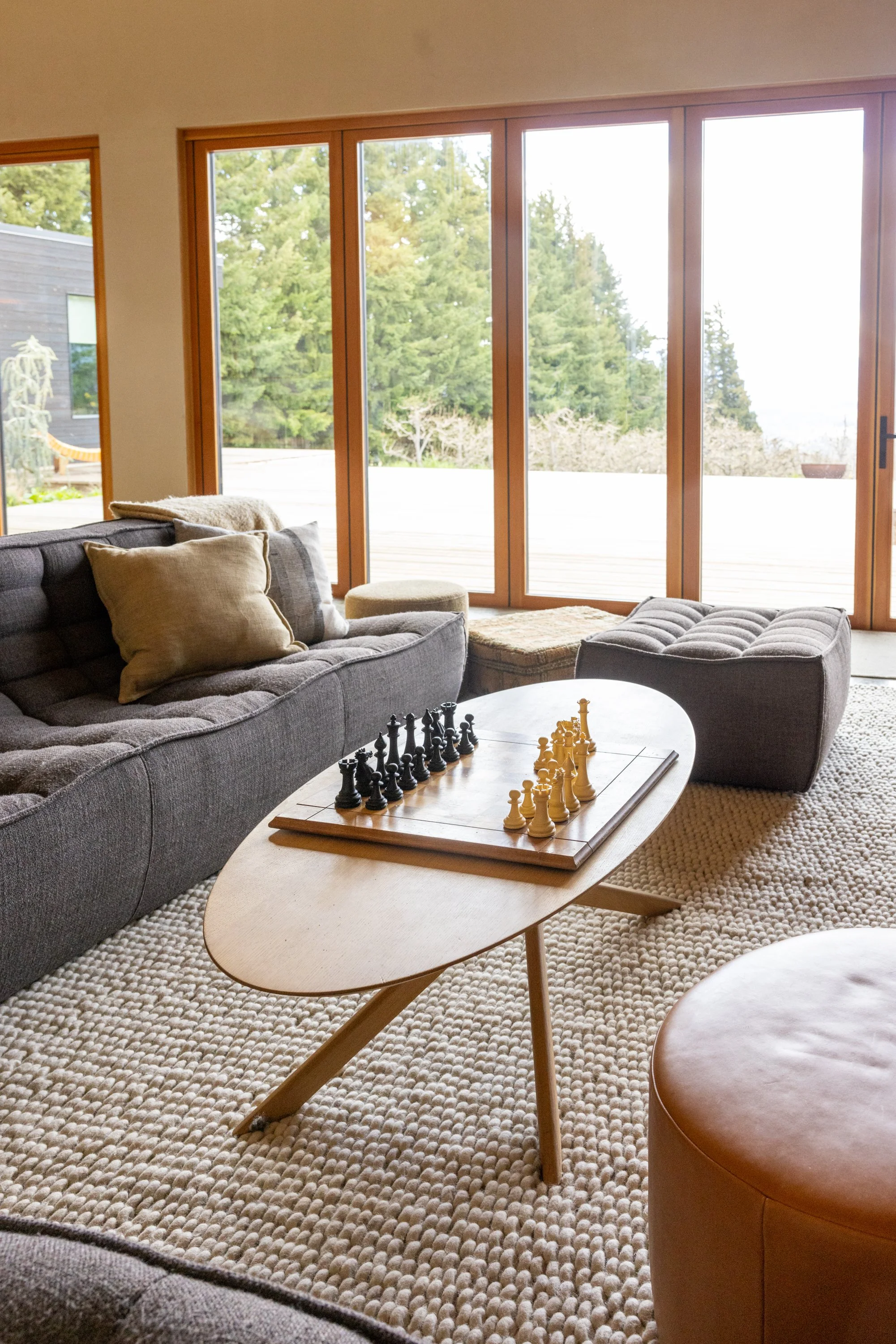 Living room with large floor-to-ceiling windows, gray couch with pillows, a round ottoman, a wooden coffee table with a chess set, and a beige textured rug.