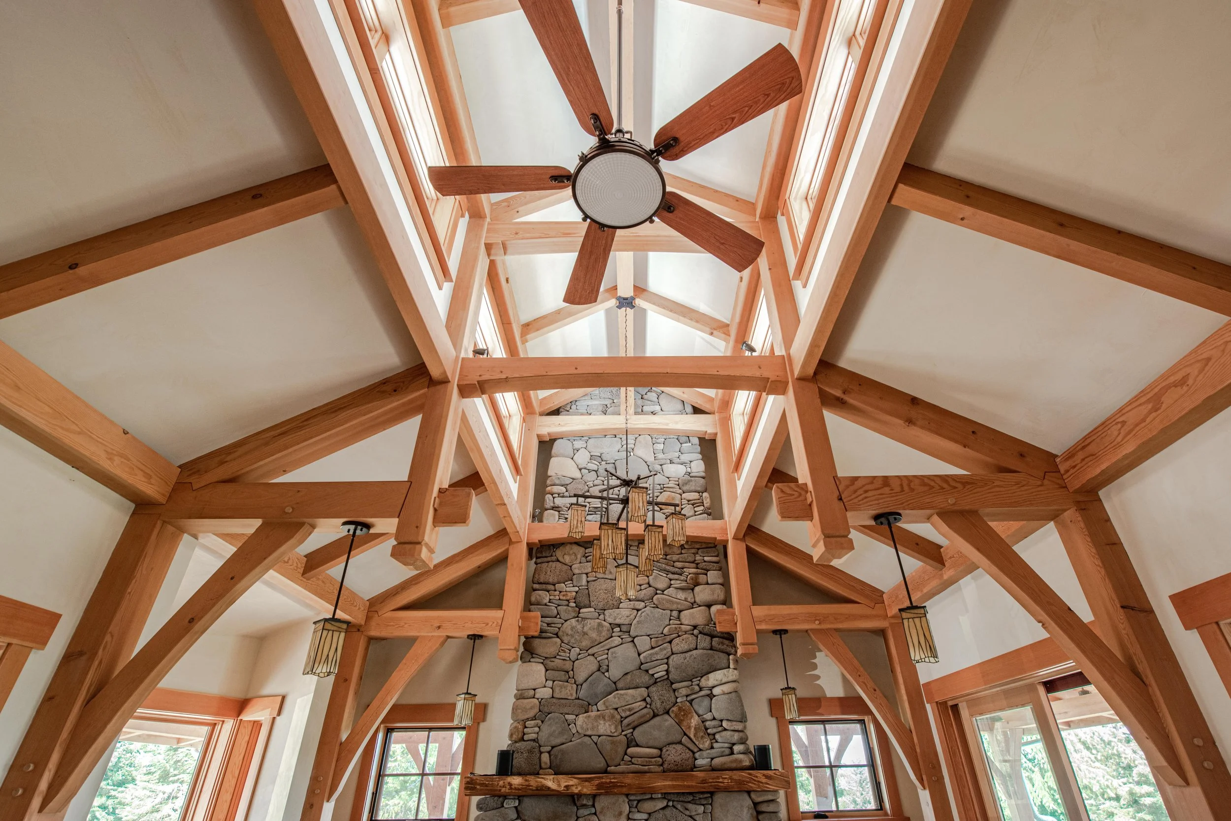 Interior view of a high ceiling with wooden beams, a ceiling fan, and large windows, centered around a stone fireplace.