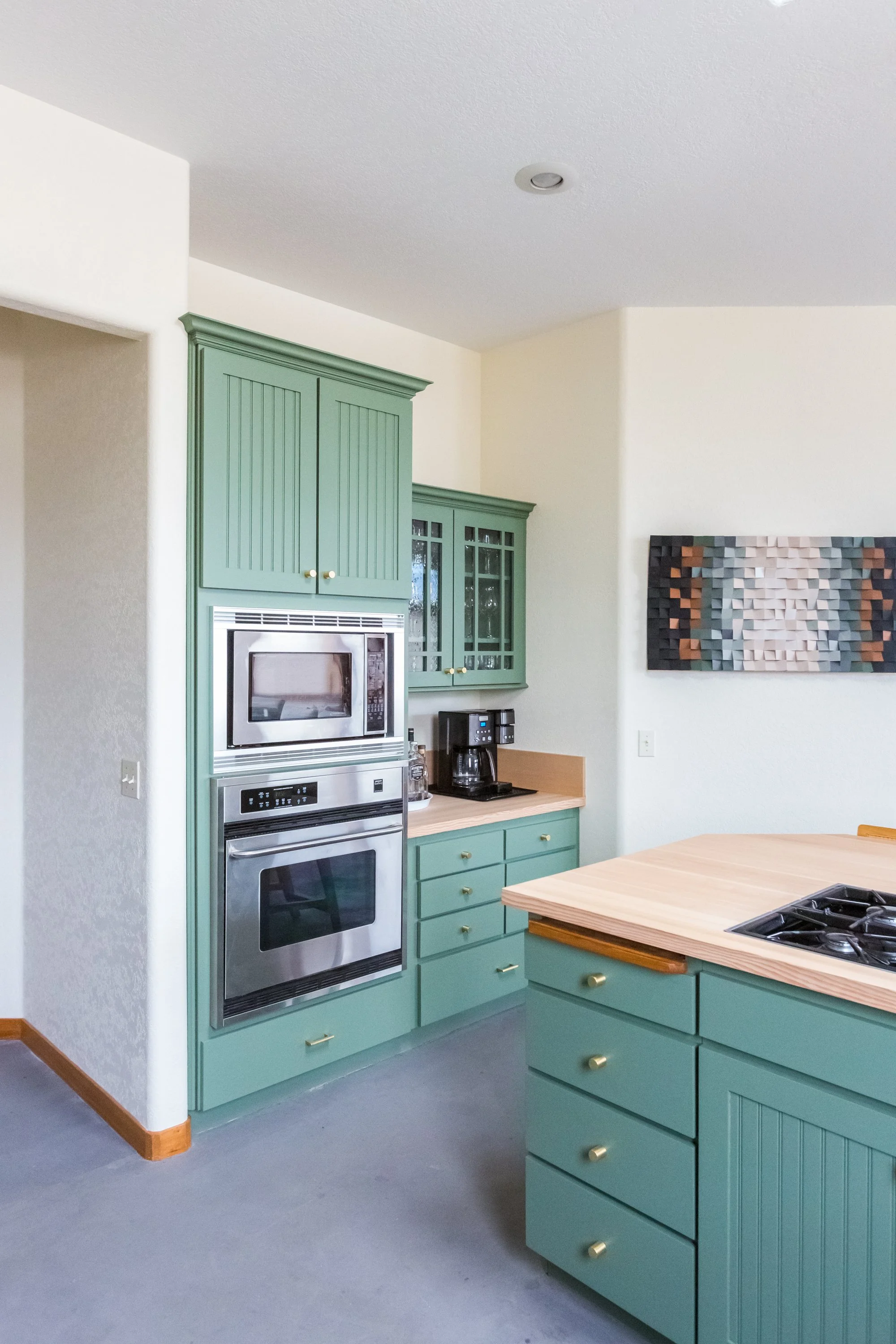 Kitchen with green cabinetry, a built-in microwave and oven, a coffee maker, and a wooden table with a stove built into the surface; artwork on the wall.