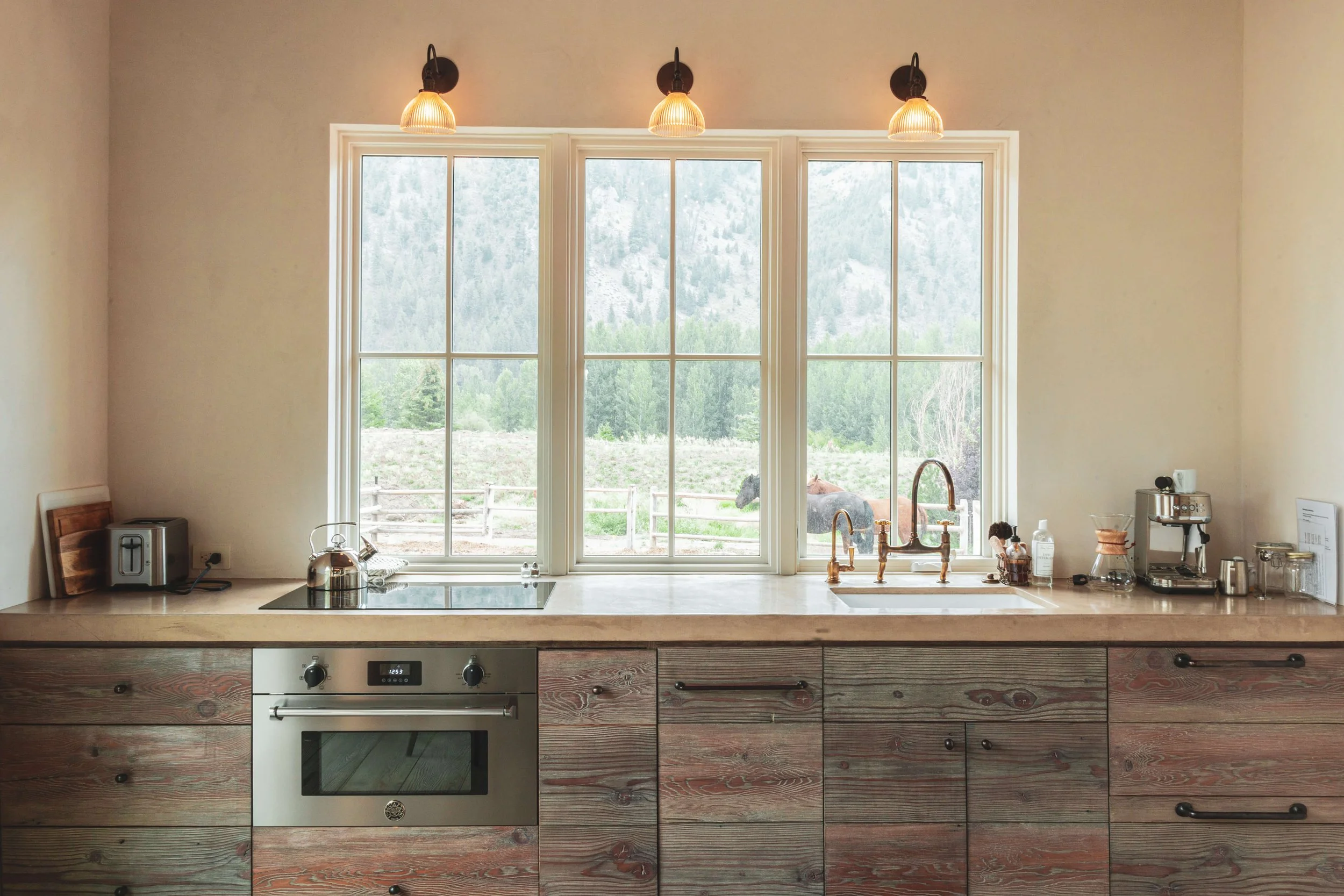 A kitchen with a large window showing a scenic view of trees and mountains outside. The countertop has a toaster, kettle, and various kitchen items near the sink. A built-in oven is below the countertop, and three light fixtures hang from the ceiling