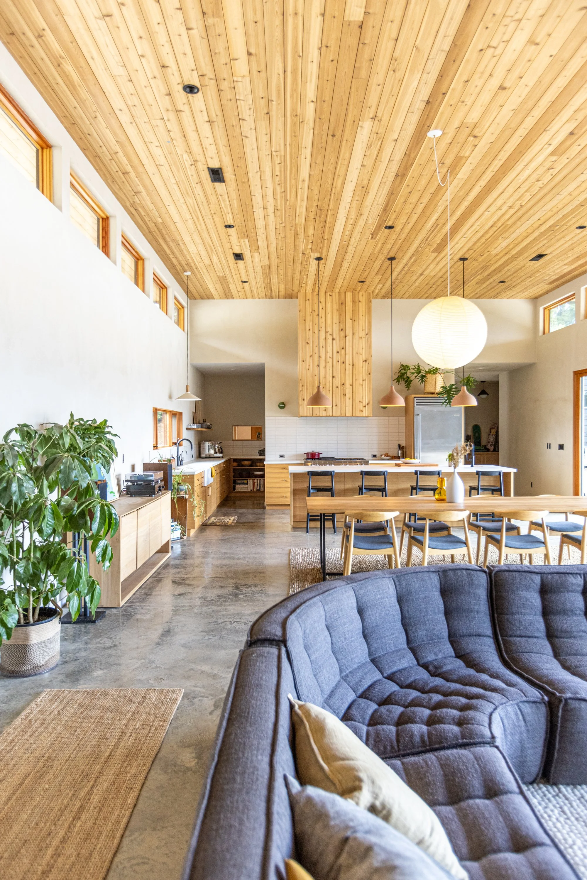 Open-concept living and kitchen area with a vaulted wooden ceiling, large windows, a gray sofa with pillows, a wooden dining table with chairs, pendant lights, and various kitchen appliances and decor.