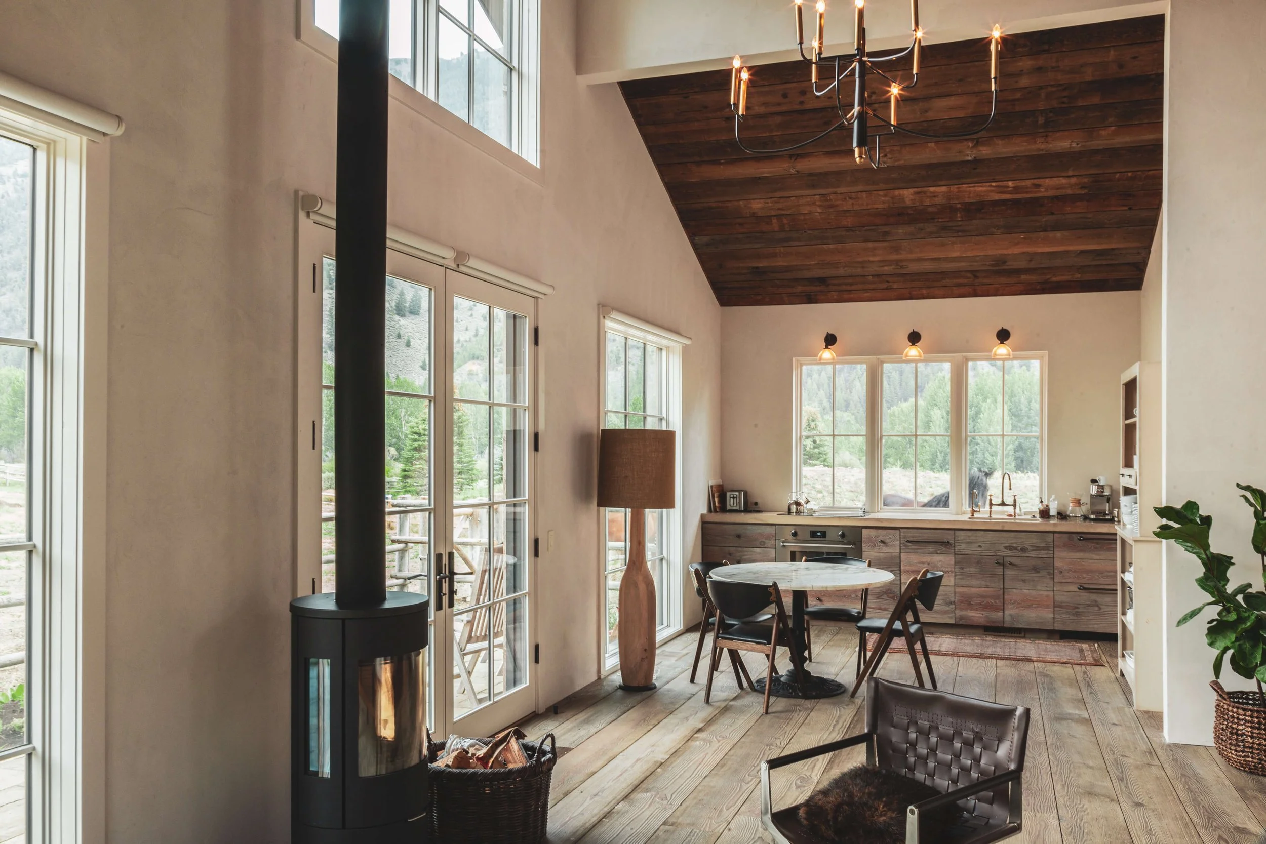 Interior of a modern kitchen and dining area with large windows, a round table with chairs, wooden cabinets, a chandelier, and a wood stove, with outdoor scenery visible through the windows.
