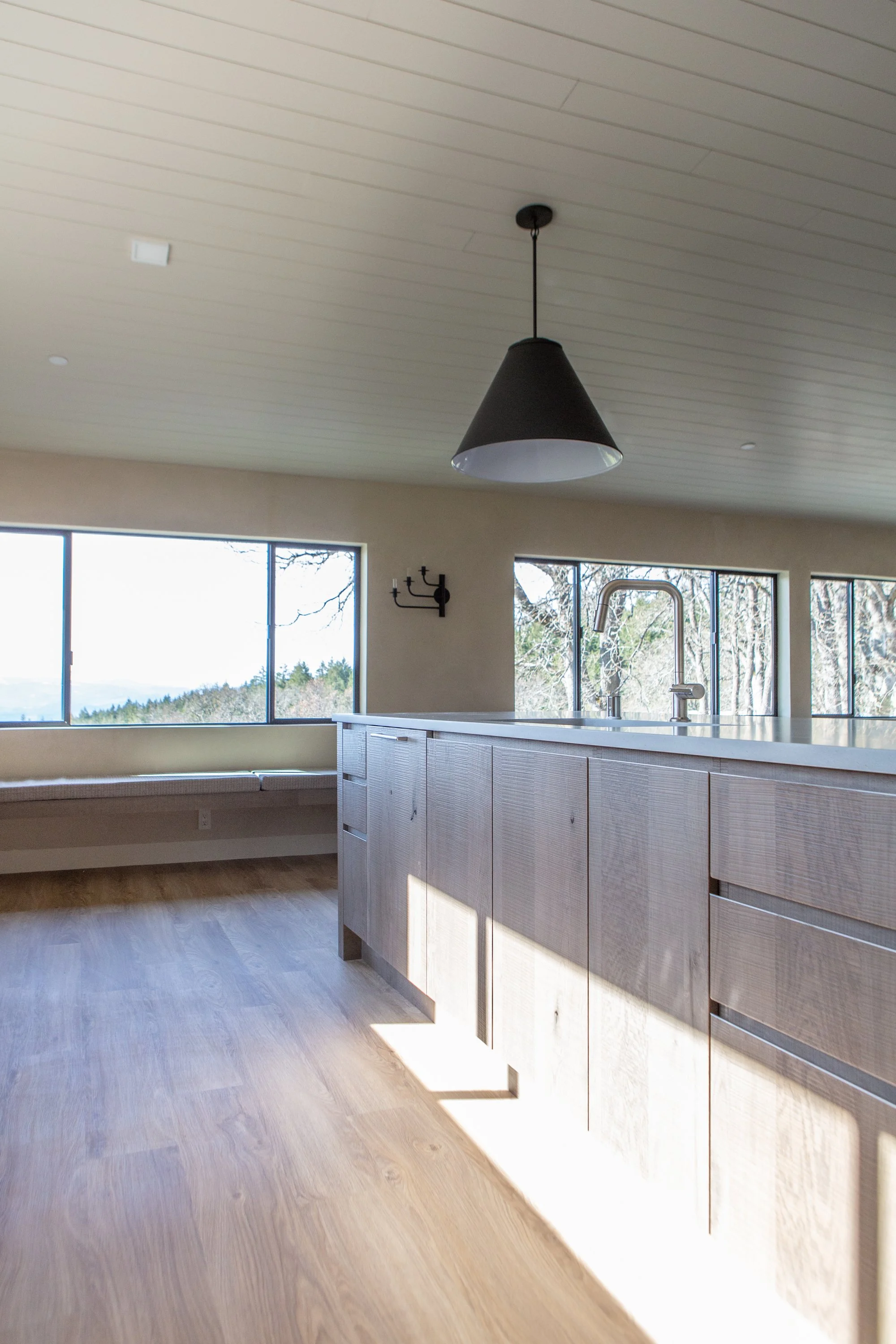 Interior view of a modern kitchen with large windows, a black pendant light hanging from the ceiling, white countertops, wooden cabinetry, and a view of trees outside.