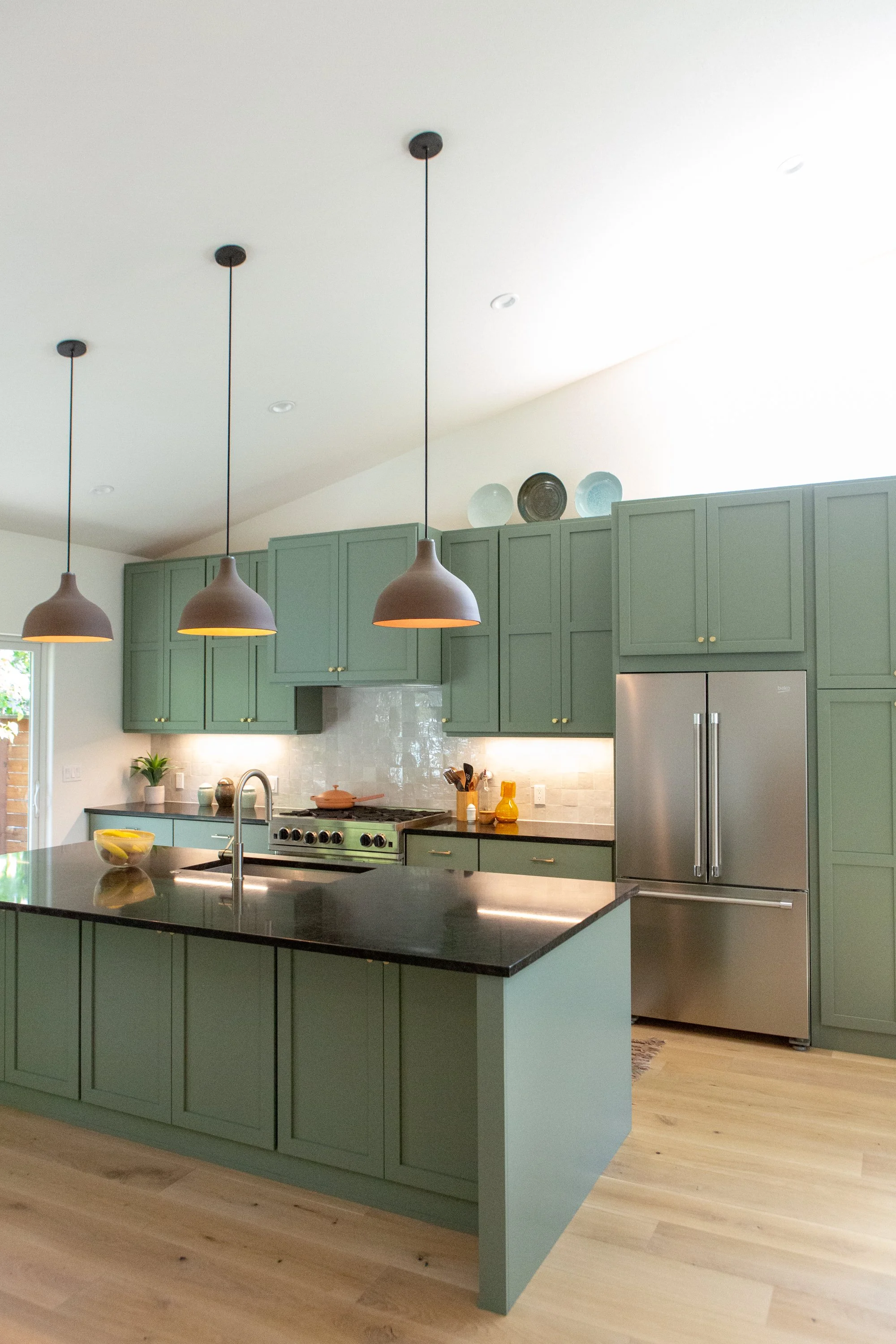 A modern kitchen with sage green cabinets, a black countertop island, pendant lighting, stainless steel appliances, and a light wood floor.