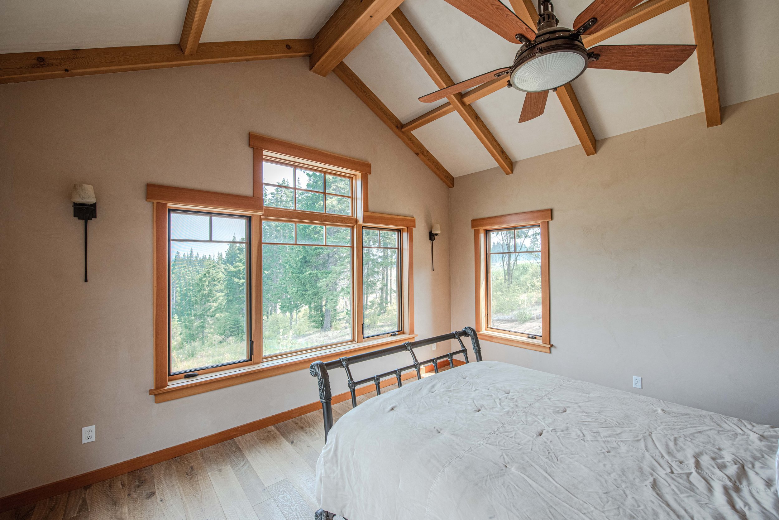 A bedroom with large windows showing a wooded landscape outside, a metal bed frame, a white mattress, ceiling wooden beams, a ceiling fan with wooden blades, and beige walls.