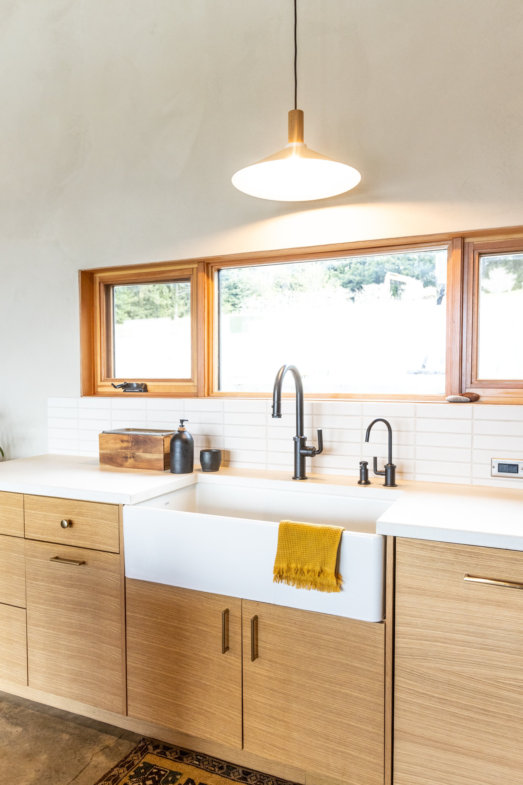 A modern kitchen with a farmhouse sink, light wood cabinets, black faucet, and three horizontal windows with wooden frames. A yellow towel hangs on the sink, and decorative black bottles sit on the counter. A hanging white and wooden pendant light il