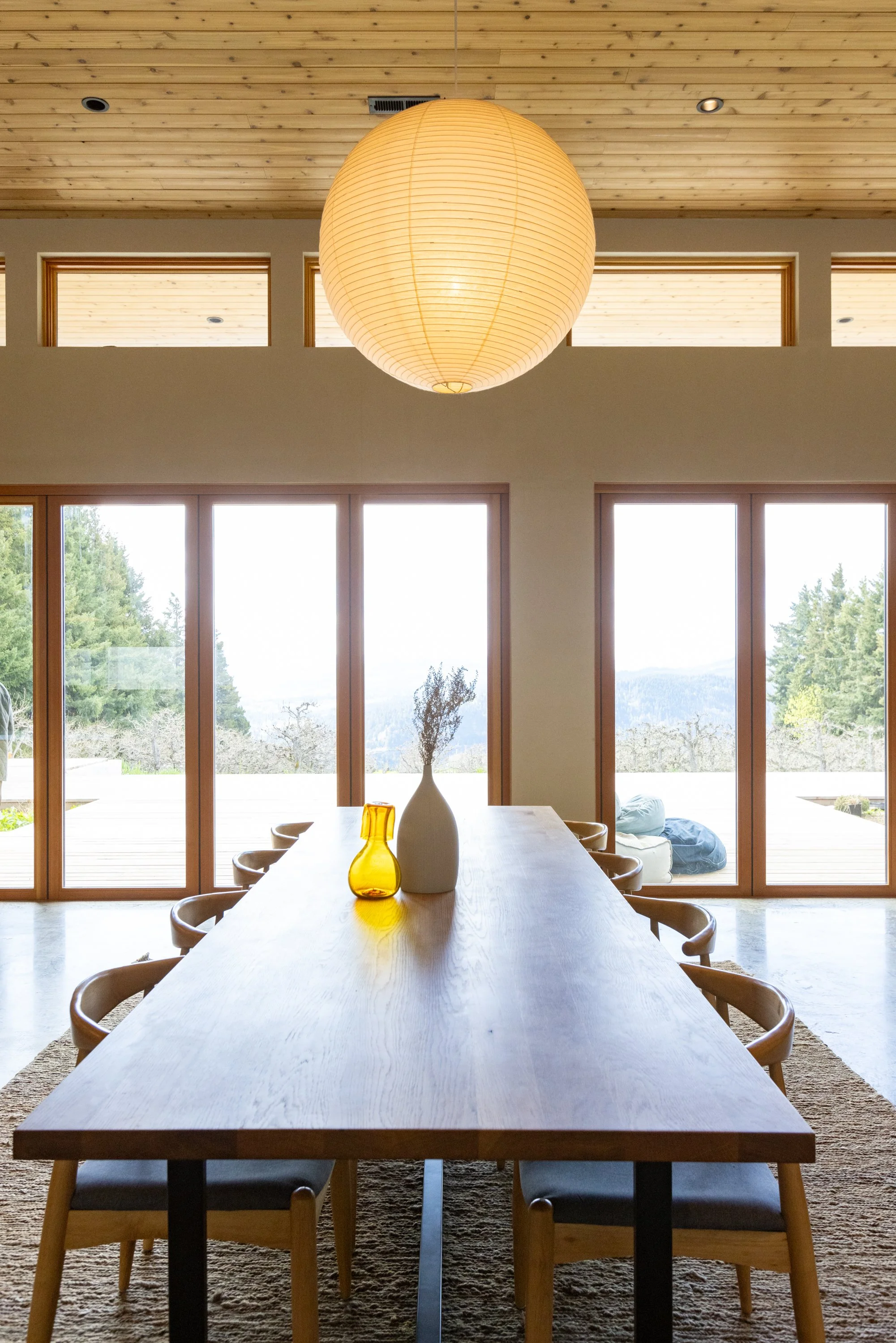 A modern dining room with a long wooden table, seating for ten, large windows showing trees and mountains outside, a large paper lantern hanging from a wooden ceiling, and decorative vases on the table.