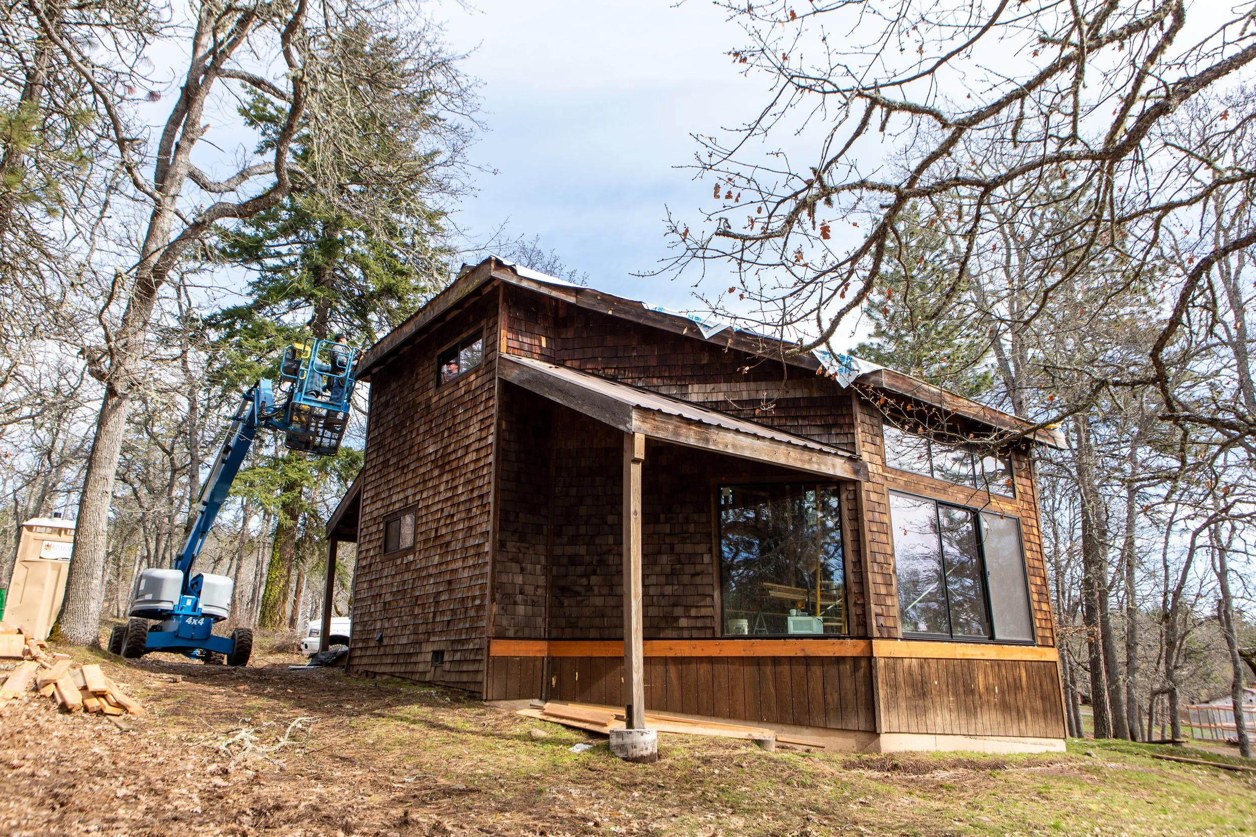A two-story wooden house with a steep roof under construction, surrounded by tall, leafless trees and a blue lift measuring workers on the roof.