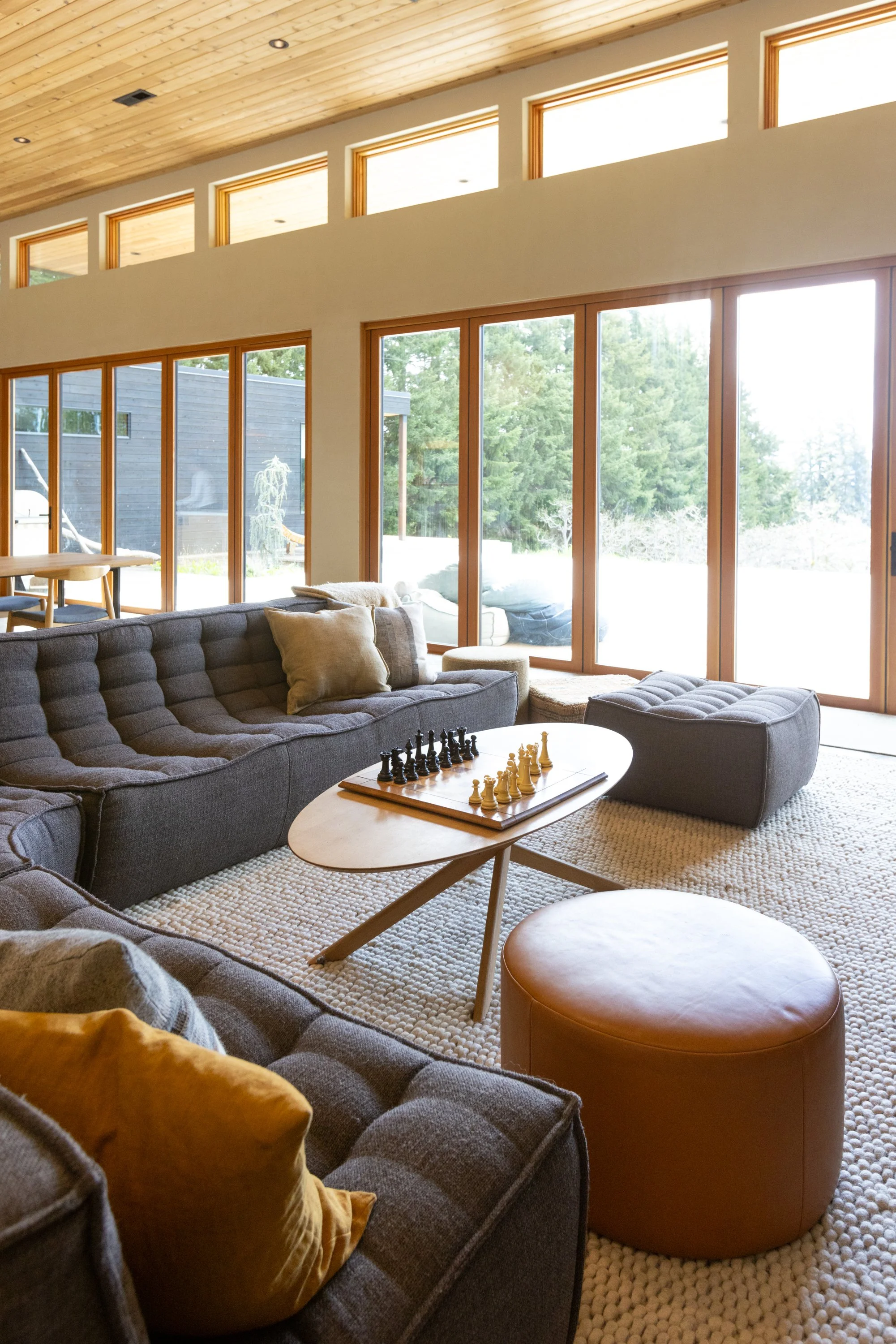 Modern living room with large glass windows, a gray sectional sofa with throw pillows, a wooden coffee table with a chessboard, and a round leather ottoman, with natural light and outdoor greenery visible.