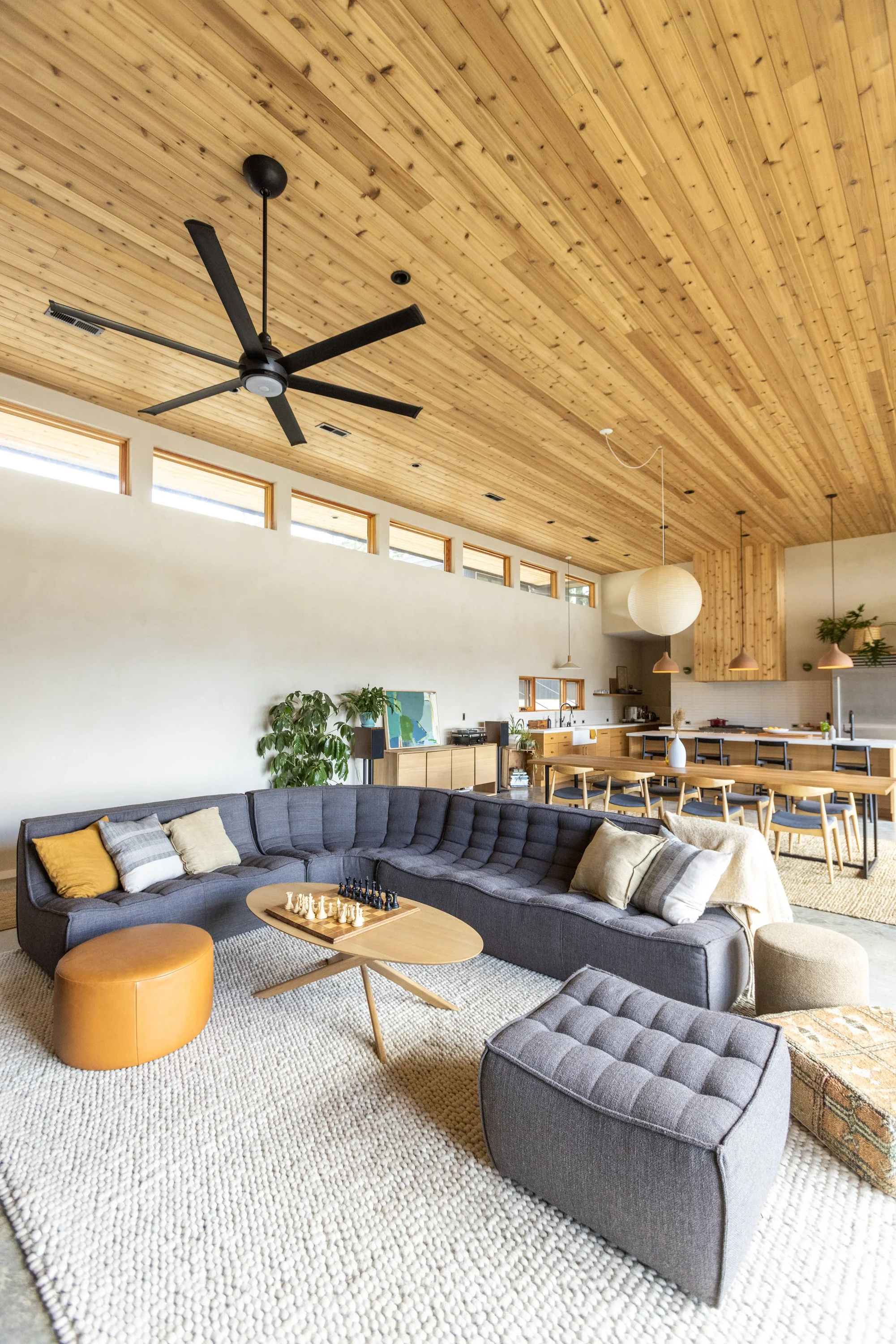 Open-concept living room with a large gray sectional sofa, coffee table with chess, mustard yellow ottoman, and white textured rug. Kitchen and dining area with wooden furniture, pendant lights, and a wooden ceiling with windows.
