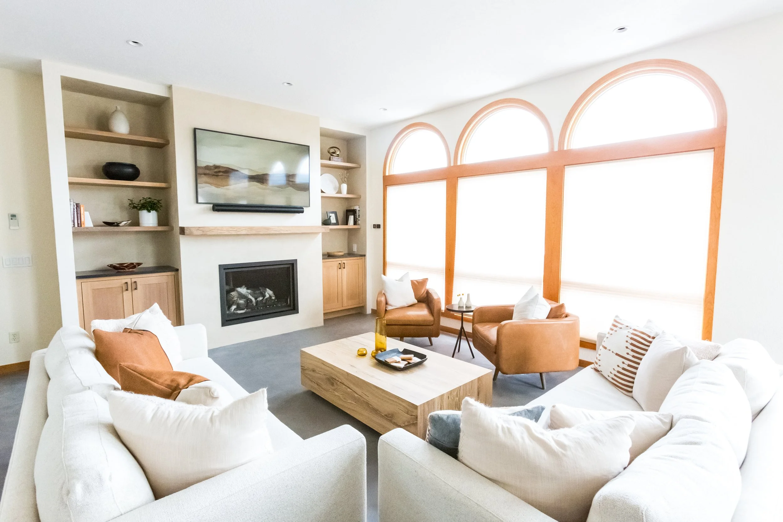 Bright living room with large arched windows, white sofa, brown leather armchairs, a light wood coffee table, and built-in shelves with decor.